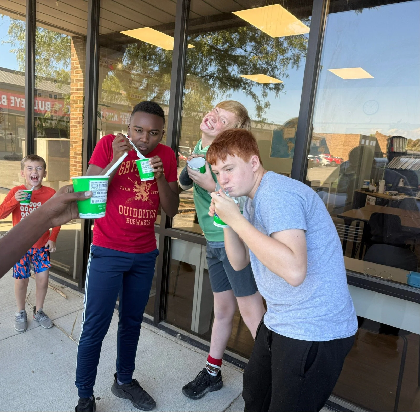 Four boys standing outside a glass building, holding cups of frozen yogurt, with one boy showing a spoonful and another with a playful expression. Two boys are in the background, one smiling and the other with an amused expression, all enjoying frozen yogurt.