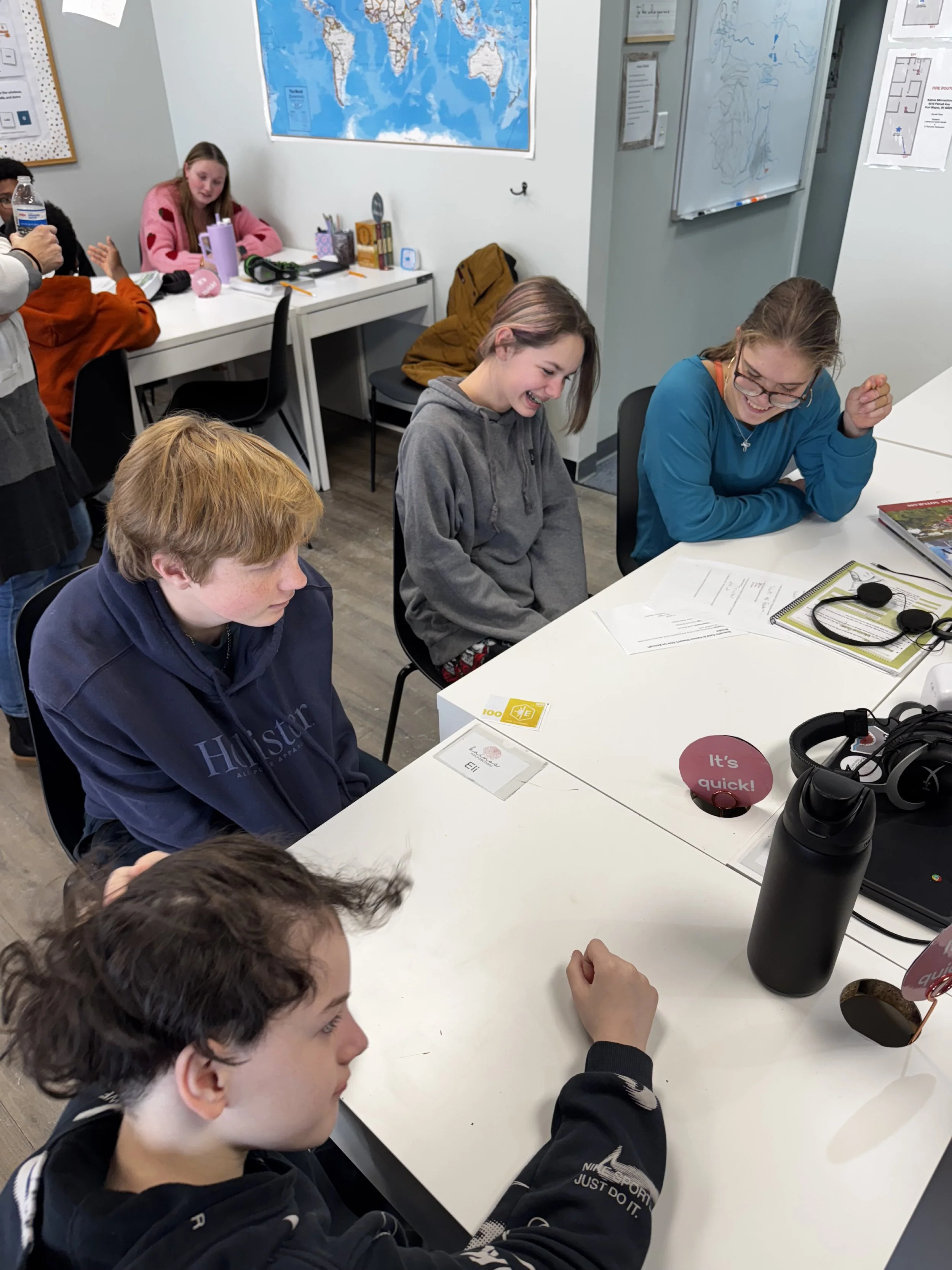 A group of students sitting and smiling around a white table in a classroom, with educational posters and a world map on the walls. Some students are looking at their papers, others are talking or laughing.