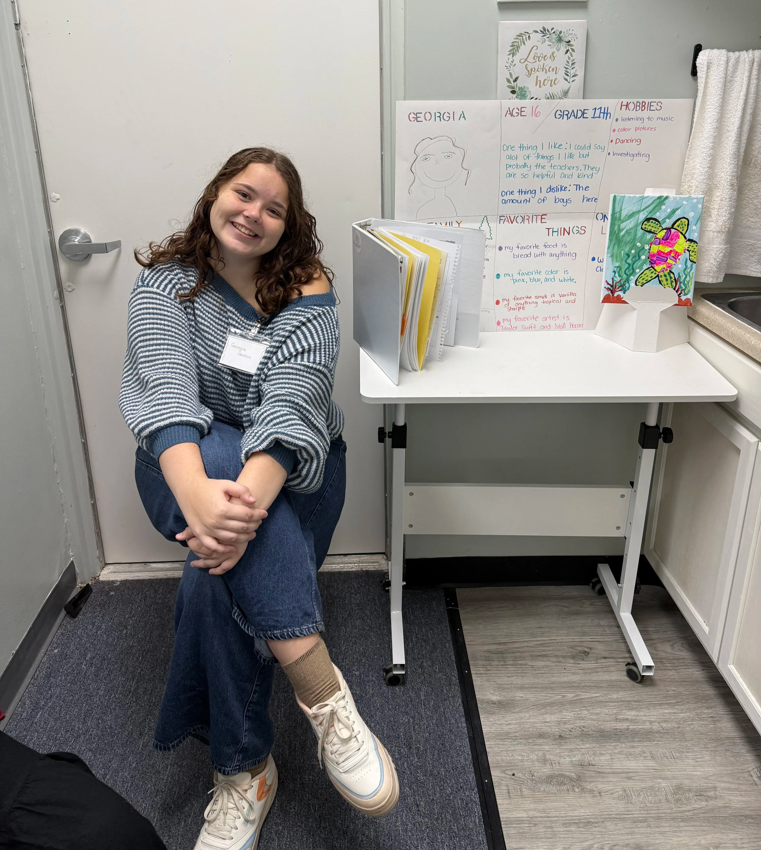 A young girl with curly brown hair smiling and posing with her arms crossed. She is wearing a blue and white striped sweater, blue jeans, knee-high socks, and white sneakers with orange accents. She is sitting in a room next to a whiteboard with handwritten notes and colorful artwork, including a frog painting on a canvas. The room has a gray door on the left, a towel hanging on a rack on the right, and a small table with papers and a folder in front of the girl.