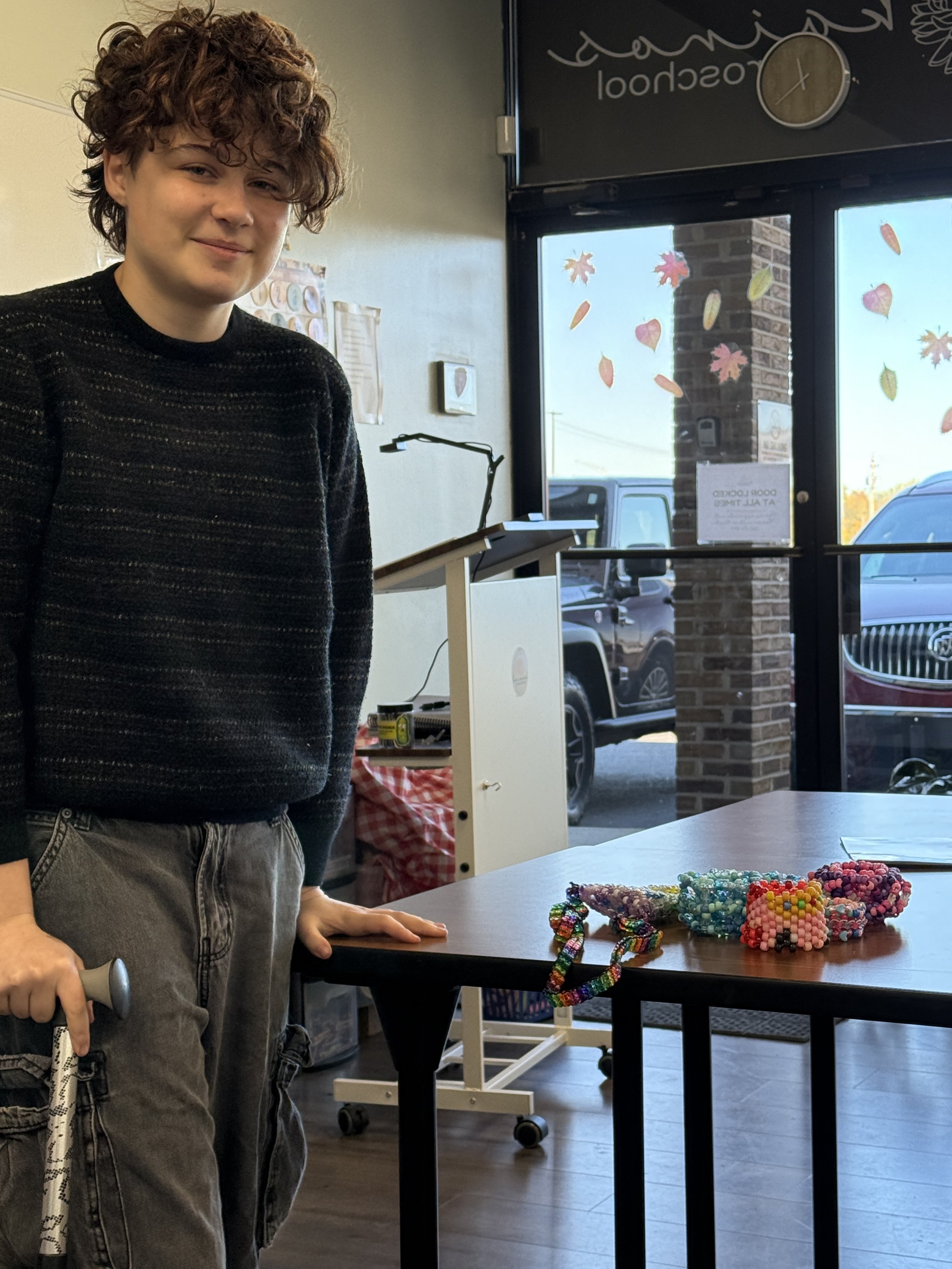A woman with short, curly hair standing next to a table decorated with colorful beaded jewelry inside a store, with a window showing parked cars outside.