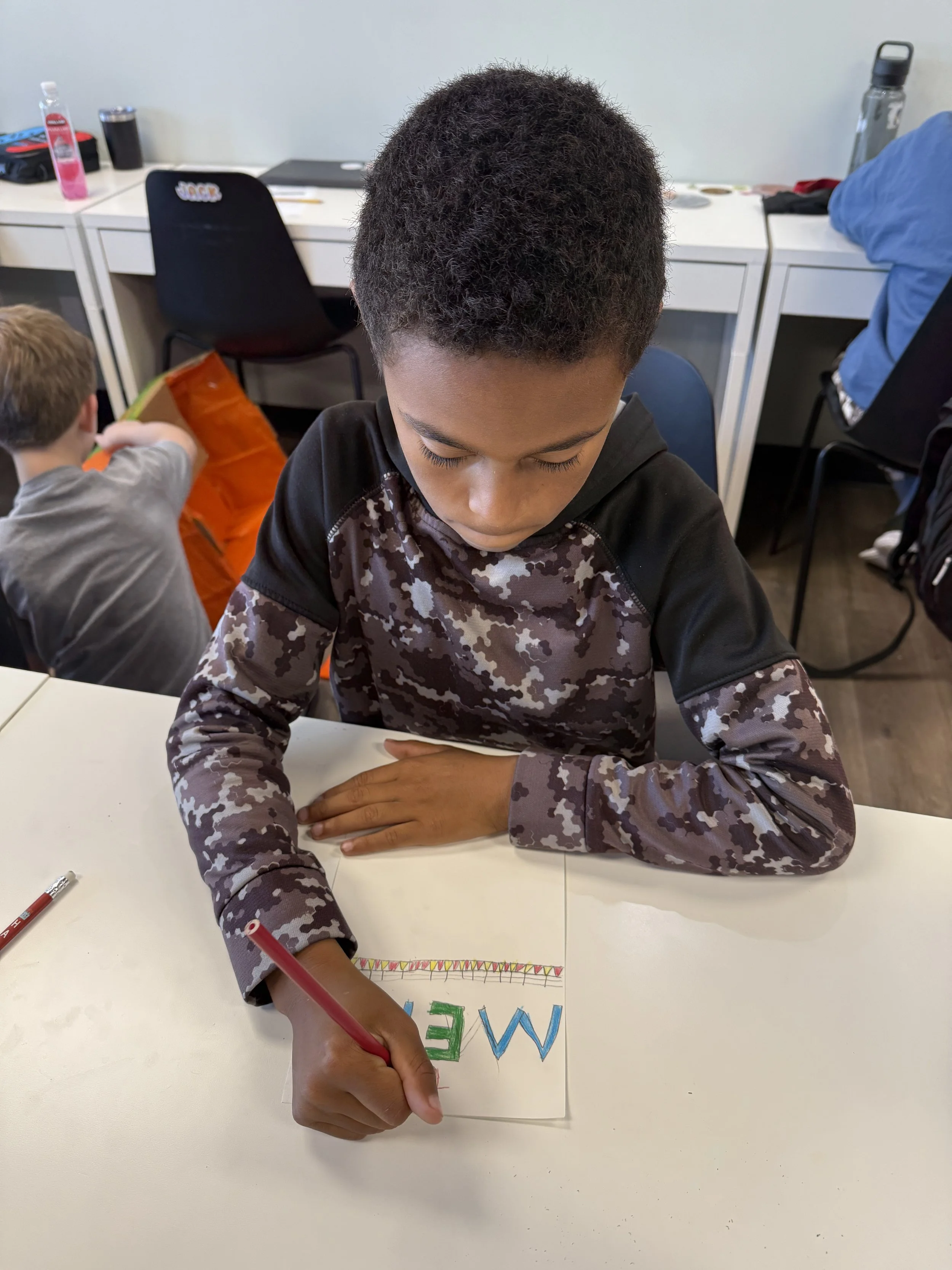 Young boy with curly hair sitting at a white table, writing on a piece of paper with colorful letters that spell 'ME' and decorations, with other children and classroom items in the background.