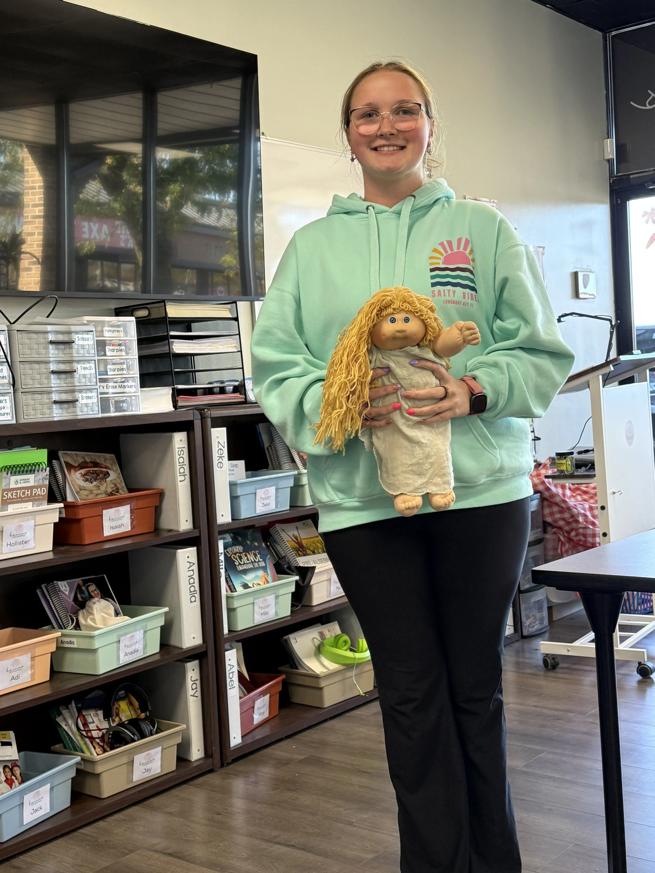 Young woman with glasses and a mint green hoodie holding a rag doll with yellow yarn hair in a classroom, surrounded by books and supplies.