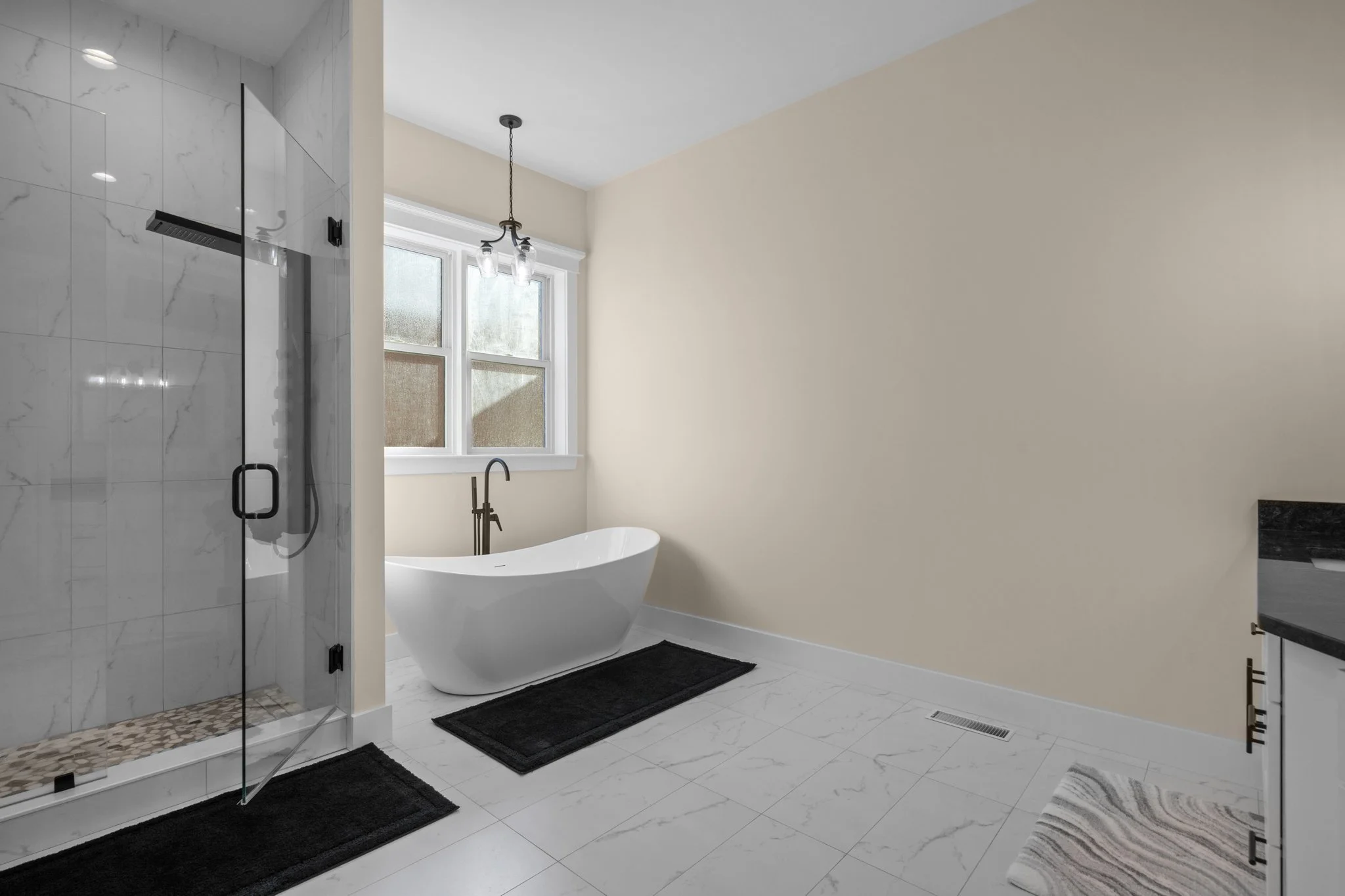 Modern bathroom with a glass-enclosed shower on the left, featuring white marble tile, a rain shower head, and pebble floor. In the background, a white freestanding bathtub under a window with frosted glass. The walls are painted light beige, with bl