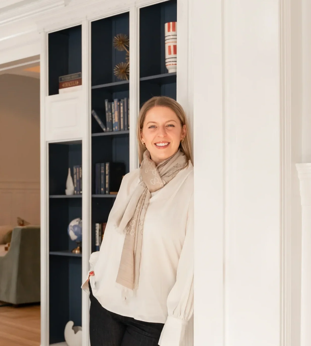 A woman with blonde hair smiling and leaning against a white wall in a living room with a bookshelf in the background.