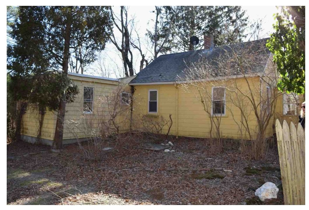Yellow house with a dark roof, surrounded by leafless shrubs and trees, with a partially visible wooden fence on the right side.
