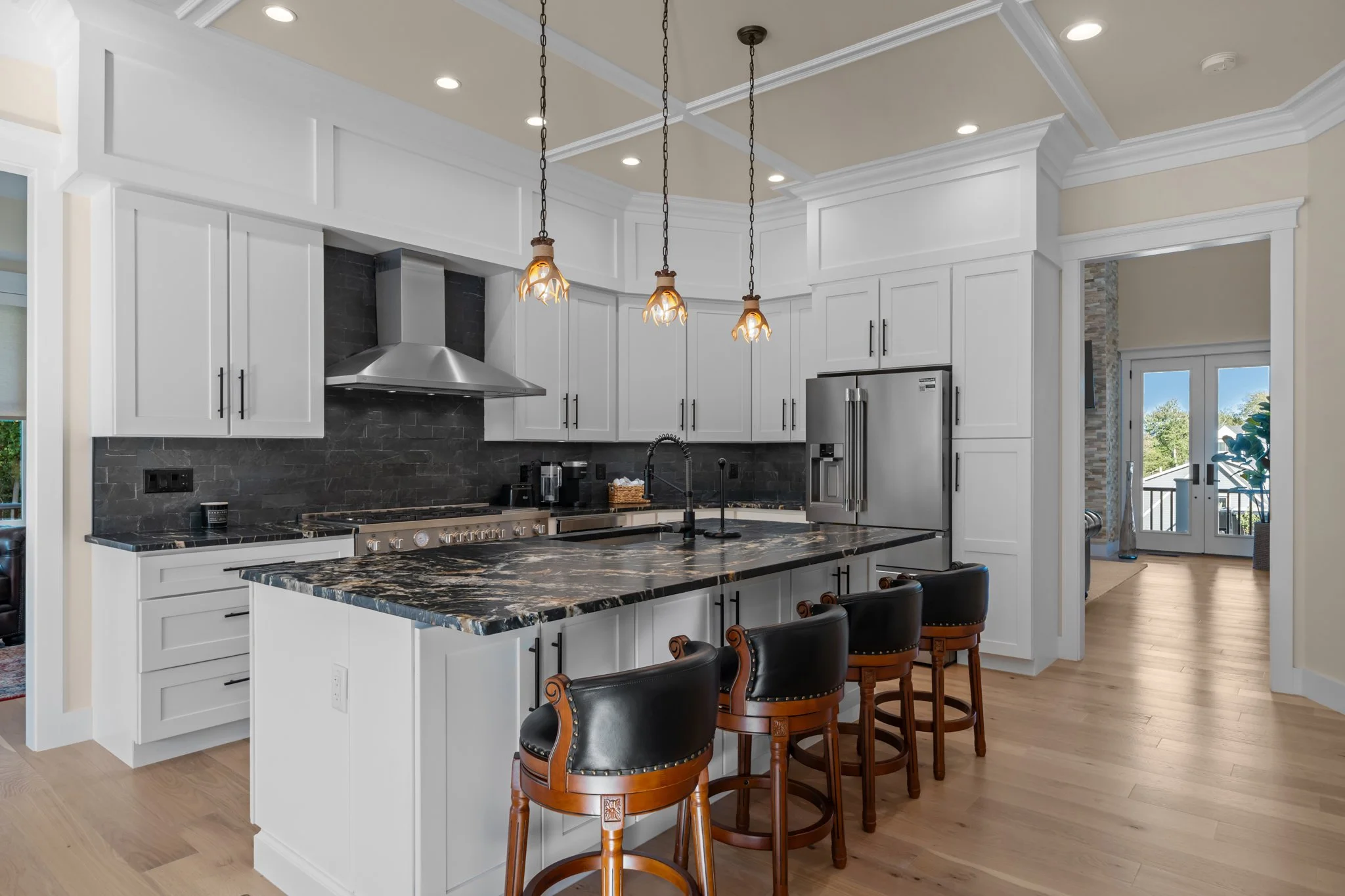 Modern kitchen with white cabinets, black stone countertops, and three pendant lights hanging above the island. There are black leather chairs around the island, and a stainless steel refrigerator next to a doorway leading outside.