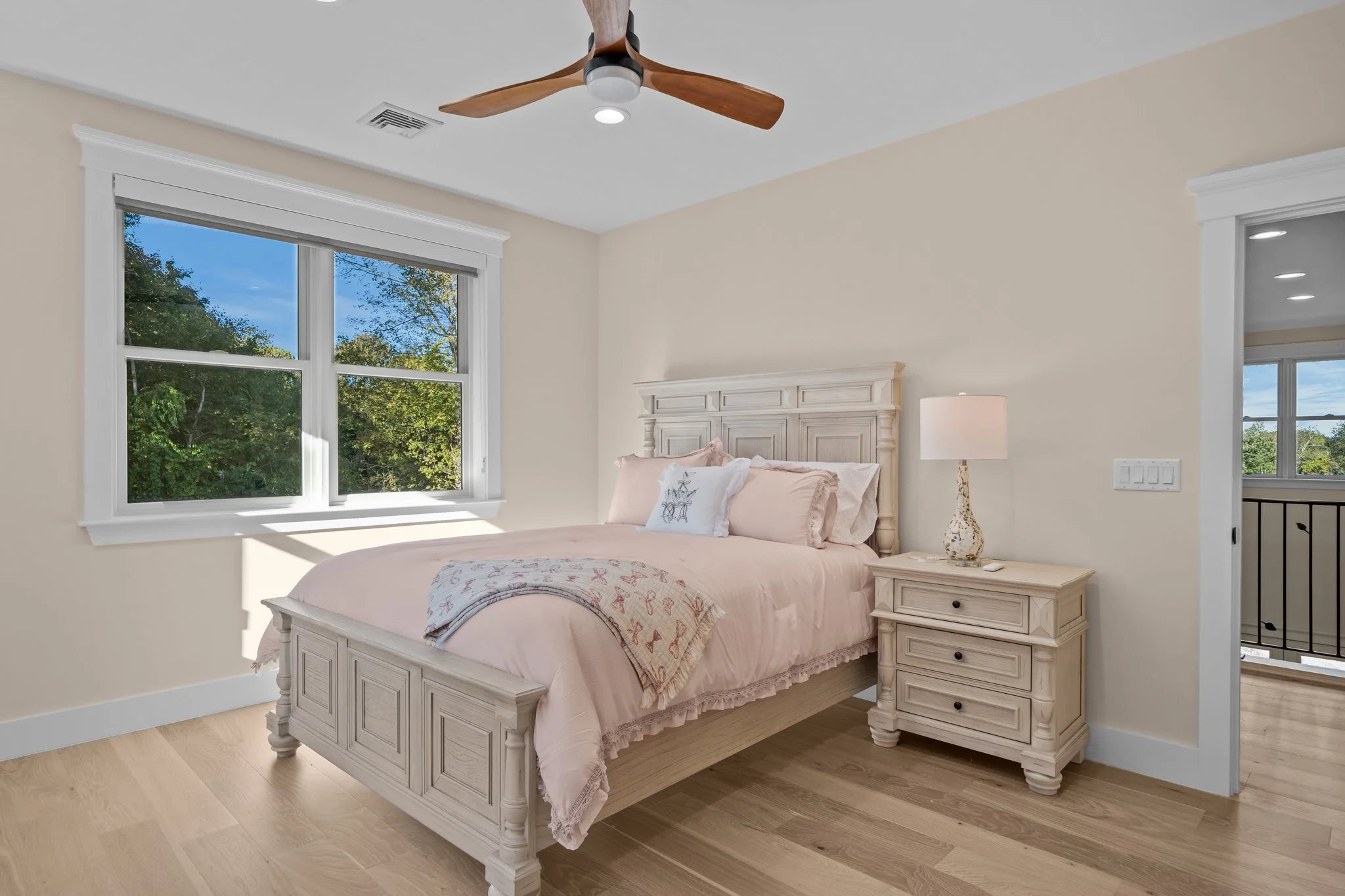 A bedroom with a large window showing trees outside. The room has a white wooden bed with a pink bedspread and pillow, a matching white nightstand with a lamp, and beige walls with white trim. A ceiling fan with a light and wooden blades is above.