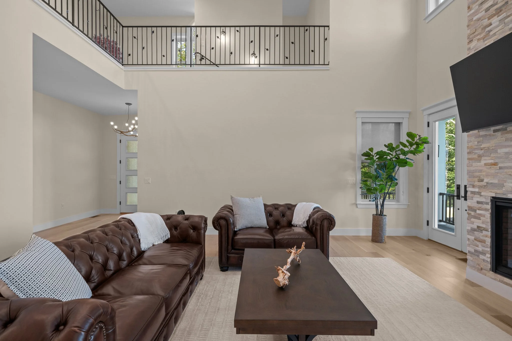 Living room with two brown leather Chesterfield sofas, a black coffee table with decor, white and black pillows, a large plant in a woven basket, stone accent wall, and large windows with view of greenery.