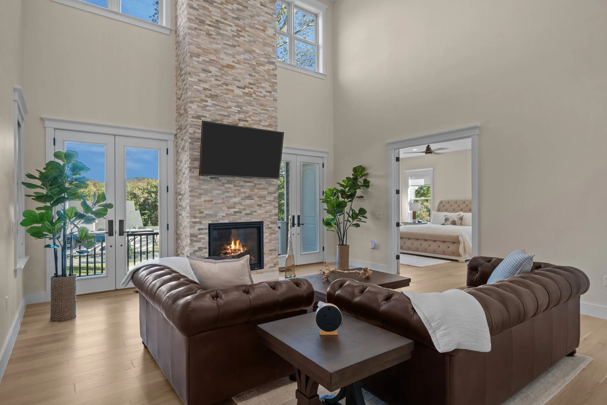 A spacious living room with a stone fireplace, a mounted flat-screen TV, large windows, and a view into a bedroom with a bed and a bedside table.