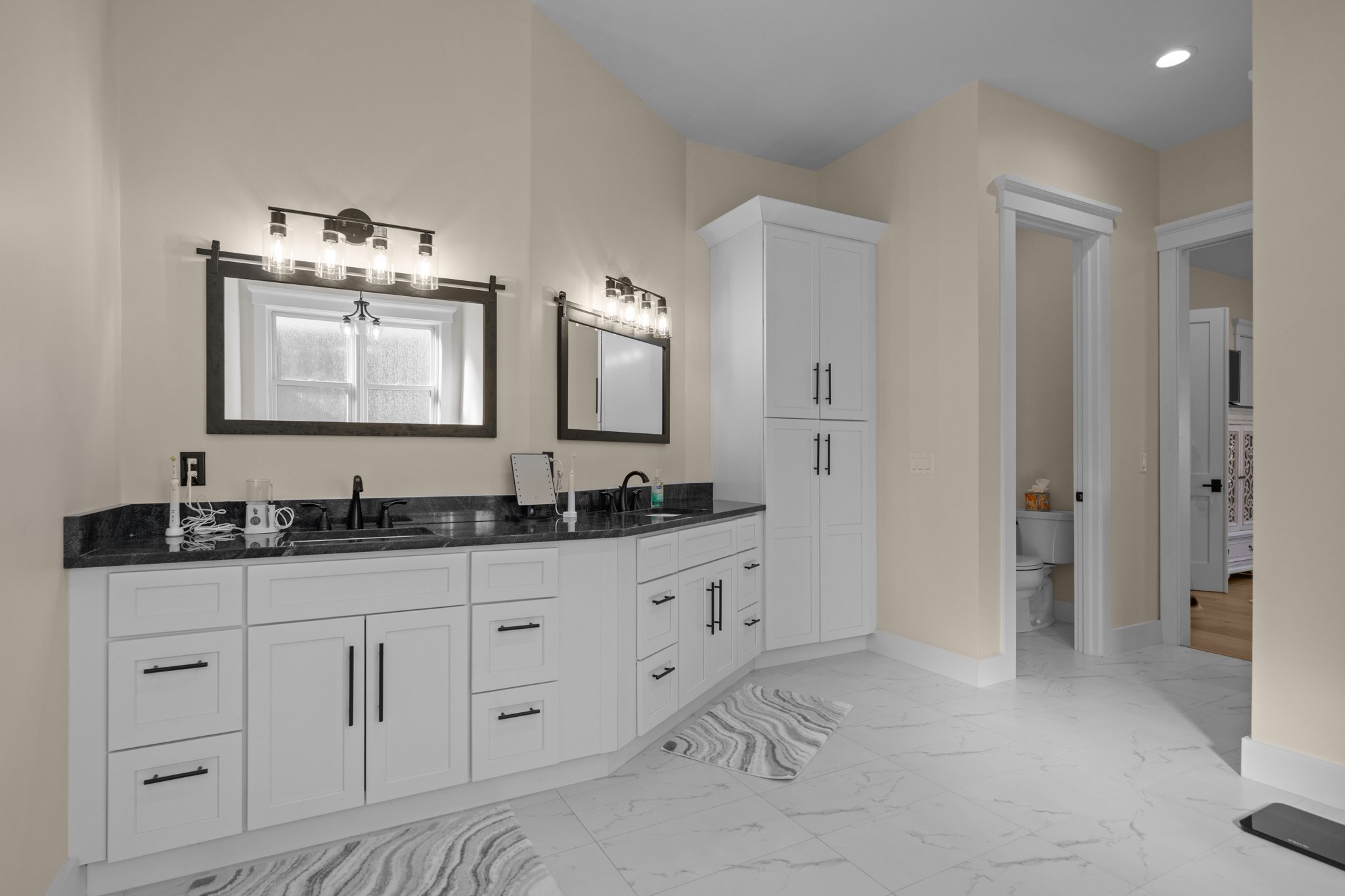 Bathroom with white cabinetry, black handles, black granite countertop, large mirrors, and lighting fixtures above the mirrors. There is a window and a small bathroom visible through a doorway, with a visible toilet.
