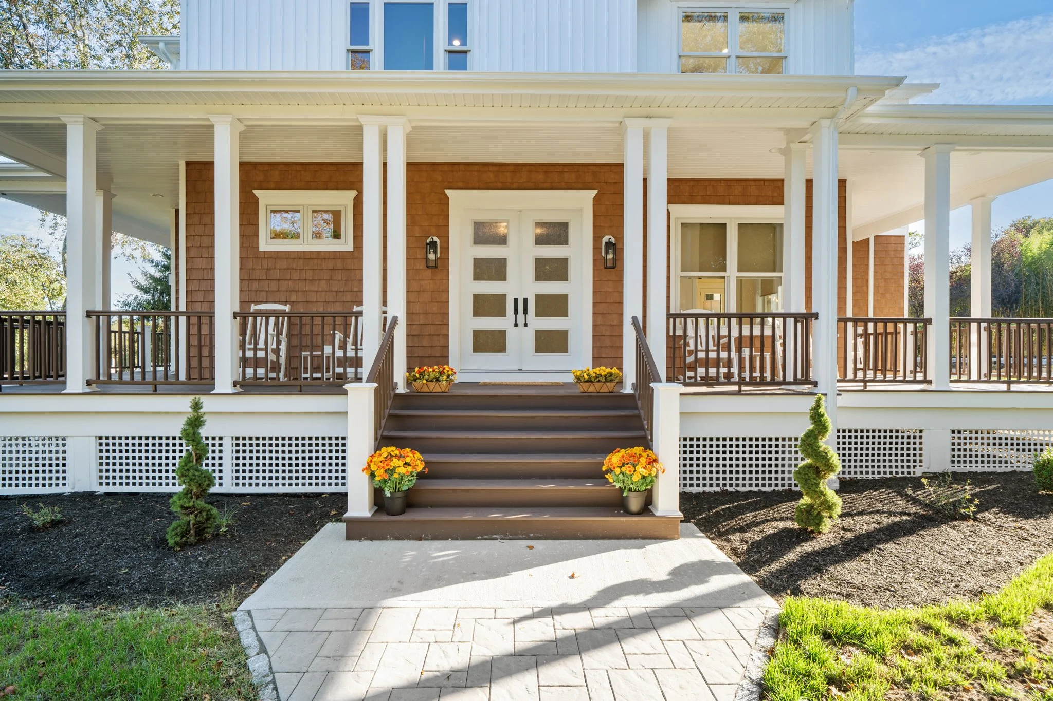 Front porch of a house with steps, potted flowers, white door, and hanging lights, surrounded by small shrubs and patches of grass.