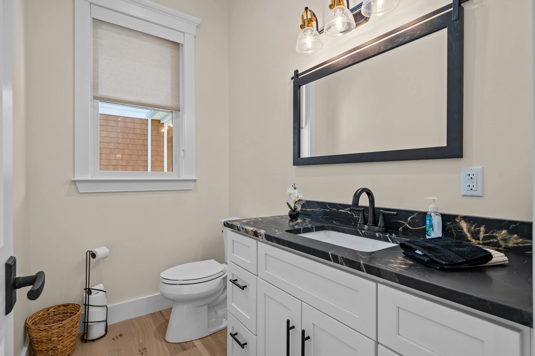 Bathroom with vanity mirror, black marble countertop, white cabinetry, toilet, window with blind, small plant, soap dispenser, towel, wicker basket, and bathroom fixtures.