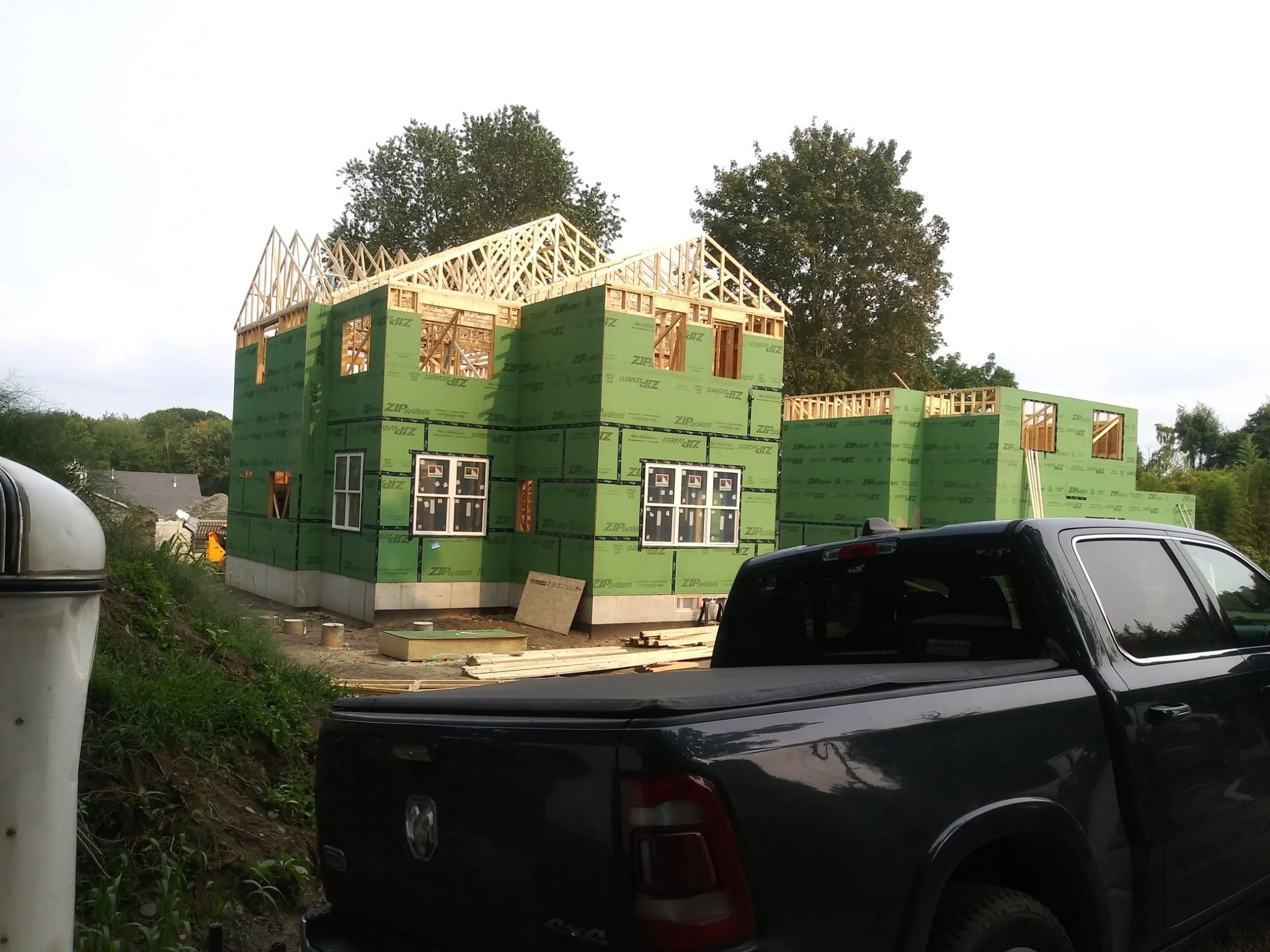 House under construction with wooden framing and green insulation panels, parked cars in the foreground, trees in the background, and a partly cloudy sky.