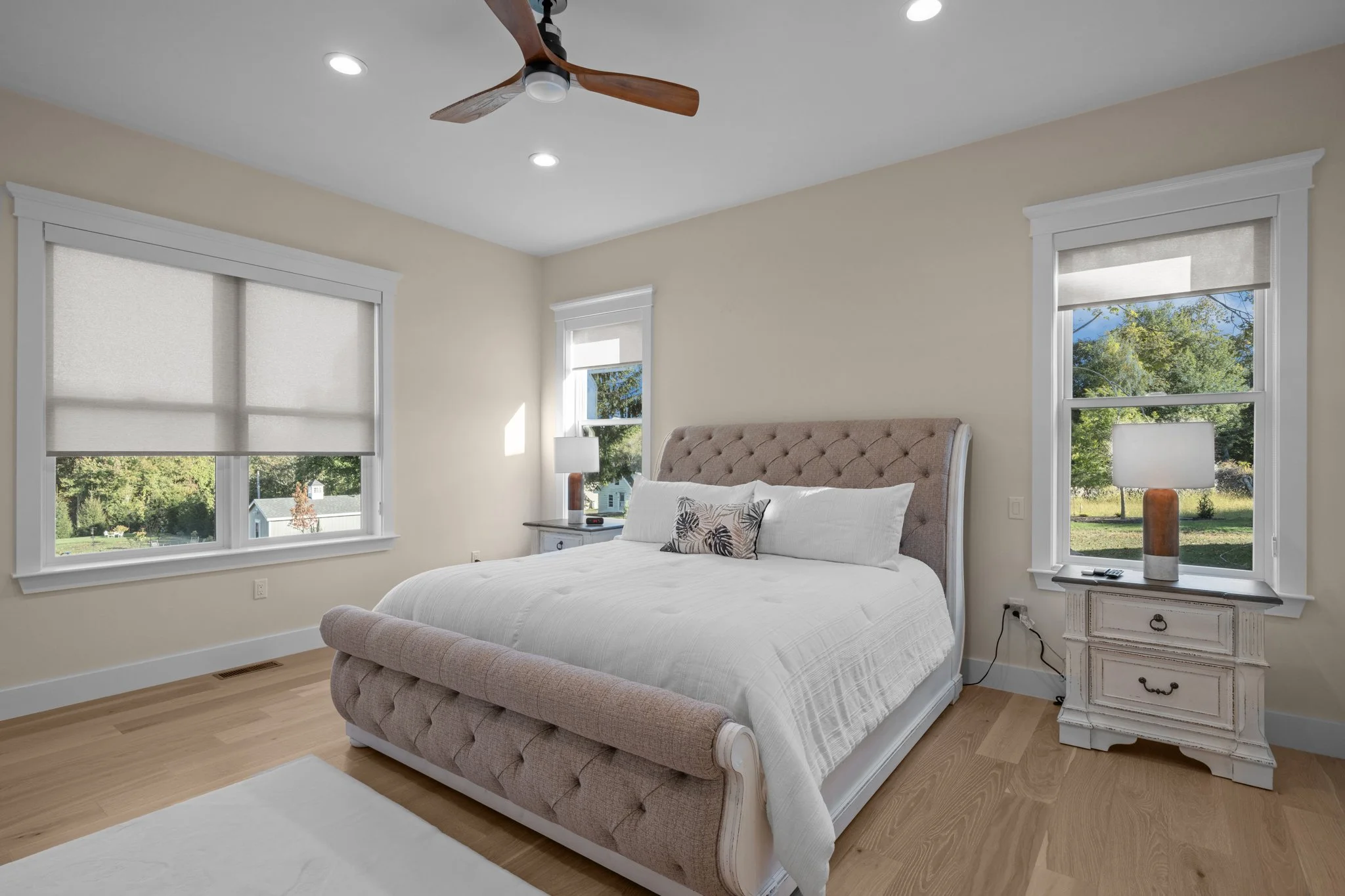 A bedroom with a bed featuring a beige tufted headboard, white bedding with decorative pillows, two white nightstands with lamps, three windows with white shades, light hardwood floors, and a ceiling fan with wooden blades.