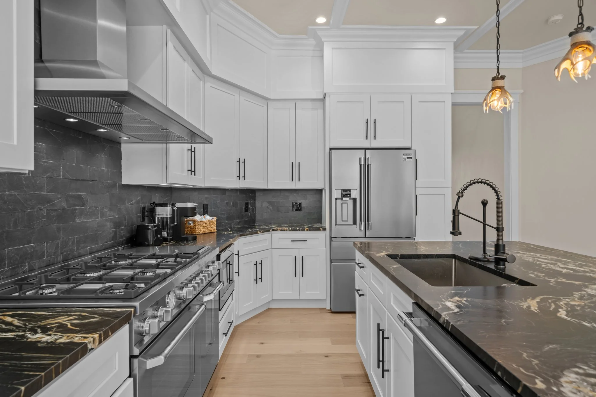 A modern kitchen with white cabinets, black hardware, and a black marble countertop with white and gold veining. There is a stainless steel refrigerator, a gas stove with multiple burners, and a black sink with a black faucet. The backsplash is made 