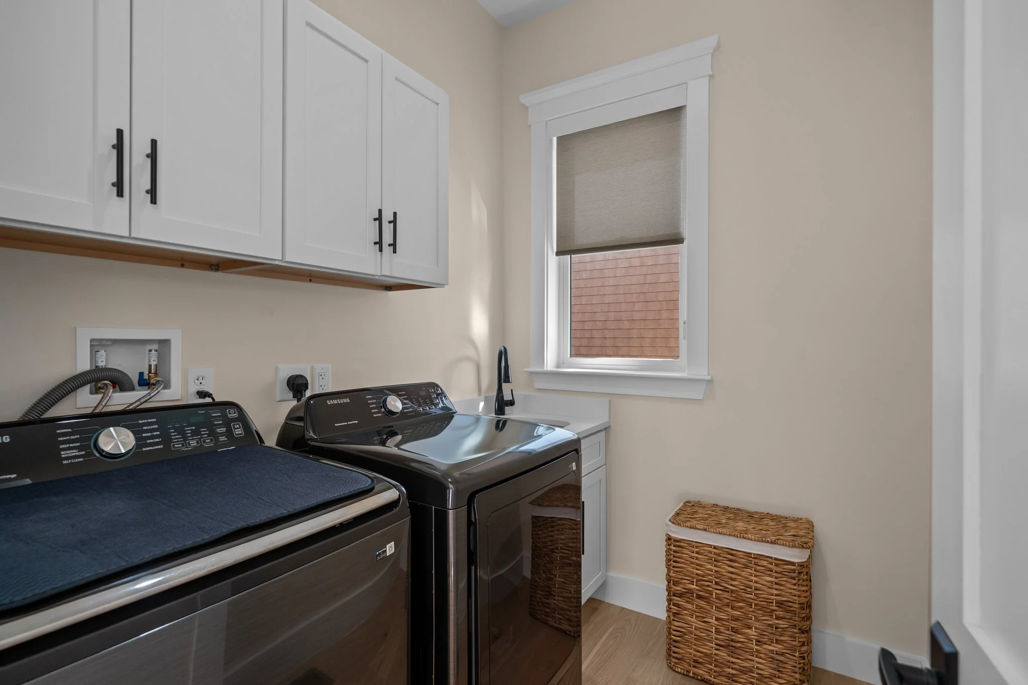 A laundry room with a white cabinet, a washer and dryer, a small window with a beige roller shade, a wicker laundry basket, and a small woven rug.