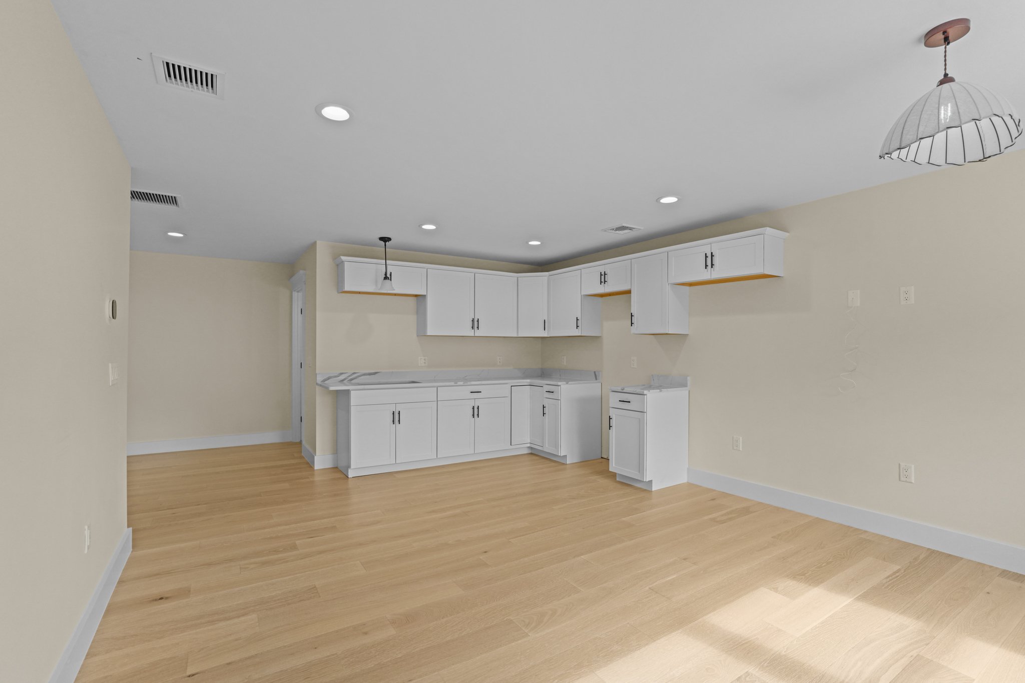 Empty kitchen with white cabinets, beige walls, wood flooring, and ceiling lights.