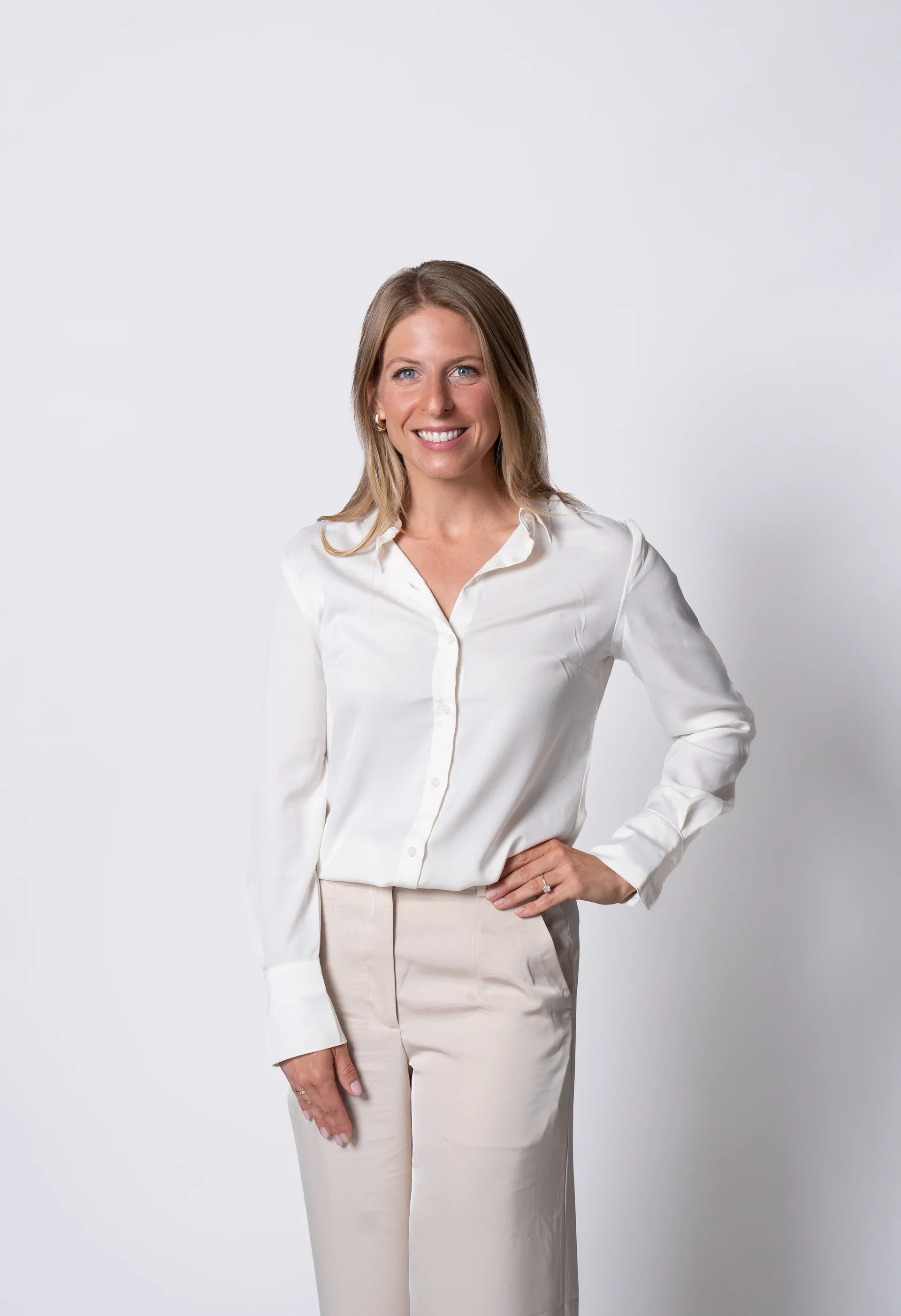 A woman with blonde hair, blue eyes, wearing a white blouse and cream-colored pants, standing against a plain white background, smiling with her right hand on her hip.