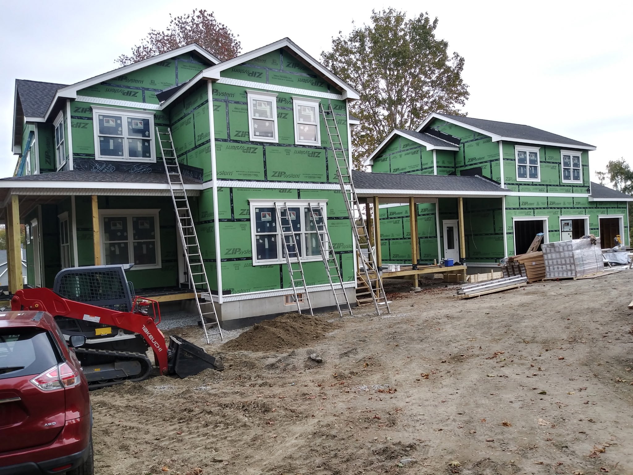 A two-story house under construction with green insulation sheathing, multiple ladders, construction materials, and a partially built porch area.