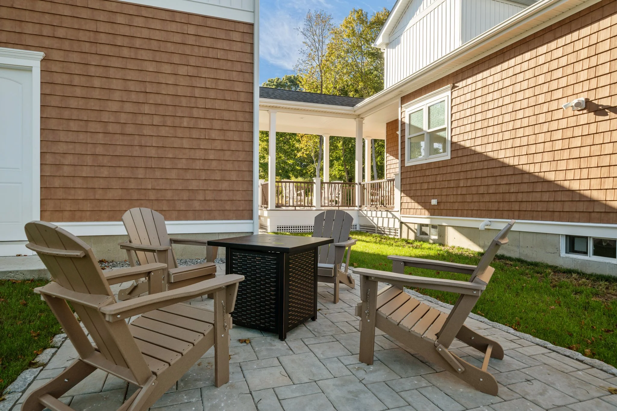 A backyard patio with multiple beige Adirondack chairs around a black fire pit, enclosed by a house with brown and white siding, and a grassy lawn with trees in the background.