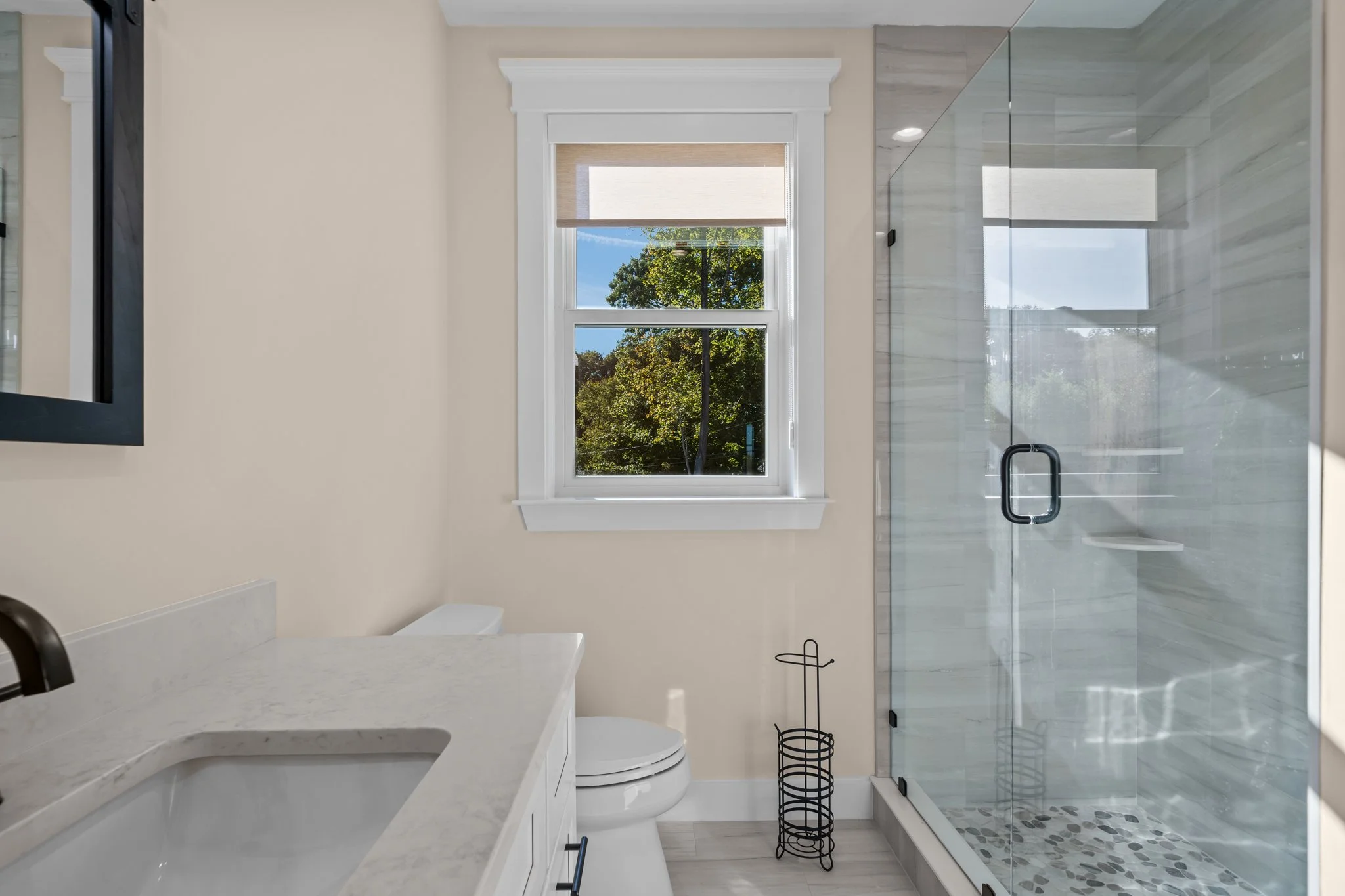Modern bathroom with a large window showing trees outside, a glass shower enclosure with black hardware, light-colored walls, and a marble countertop.
