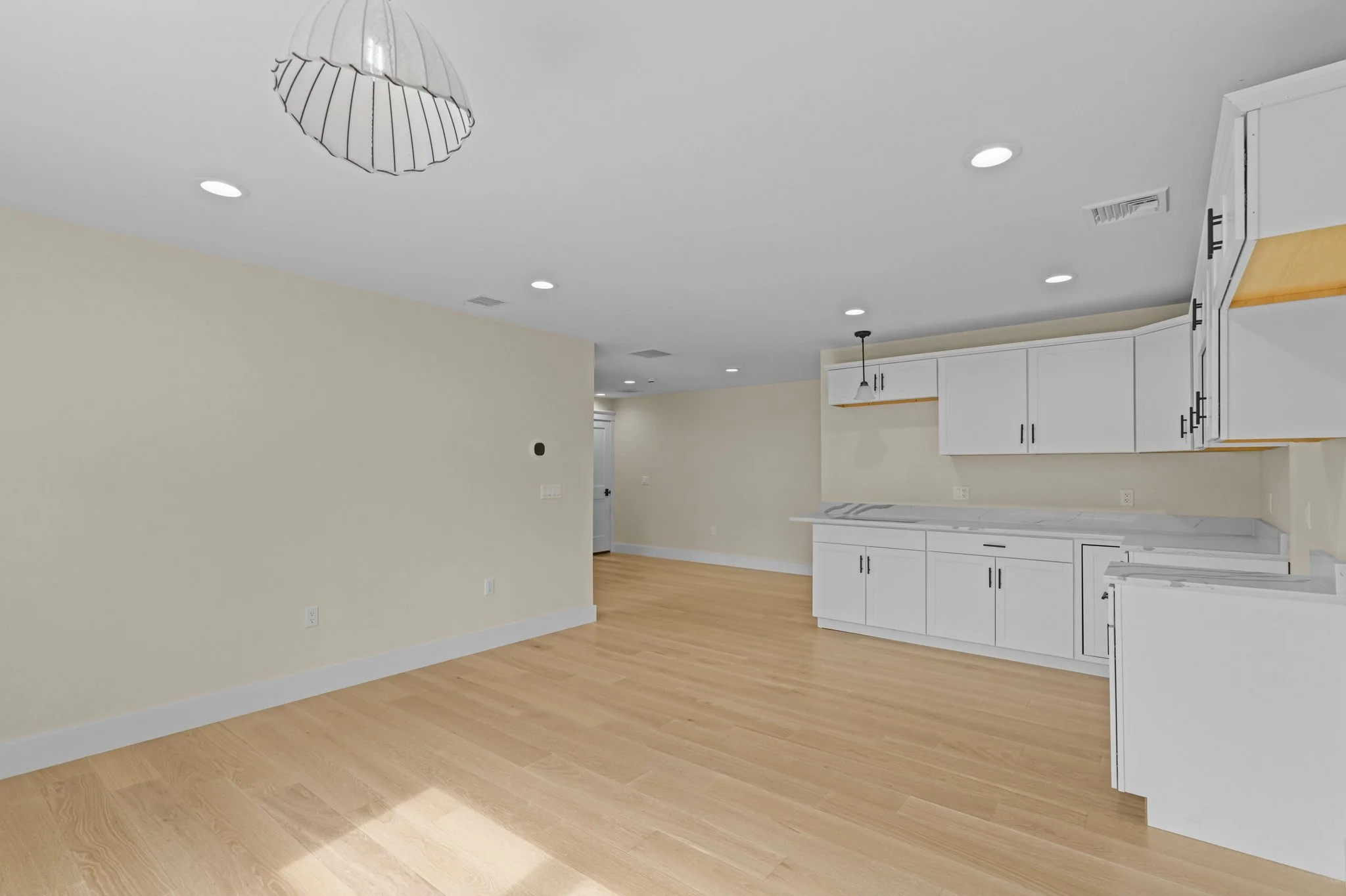 Empty kitchen and living space with wooden flooring, white cabinets, ceiling lights, and a hanging light fixture.