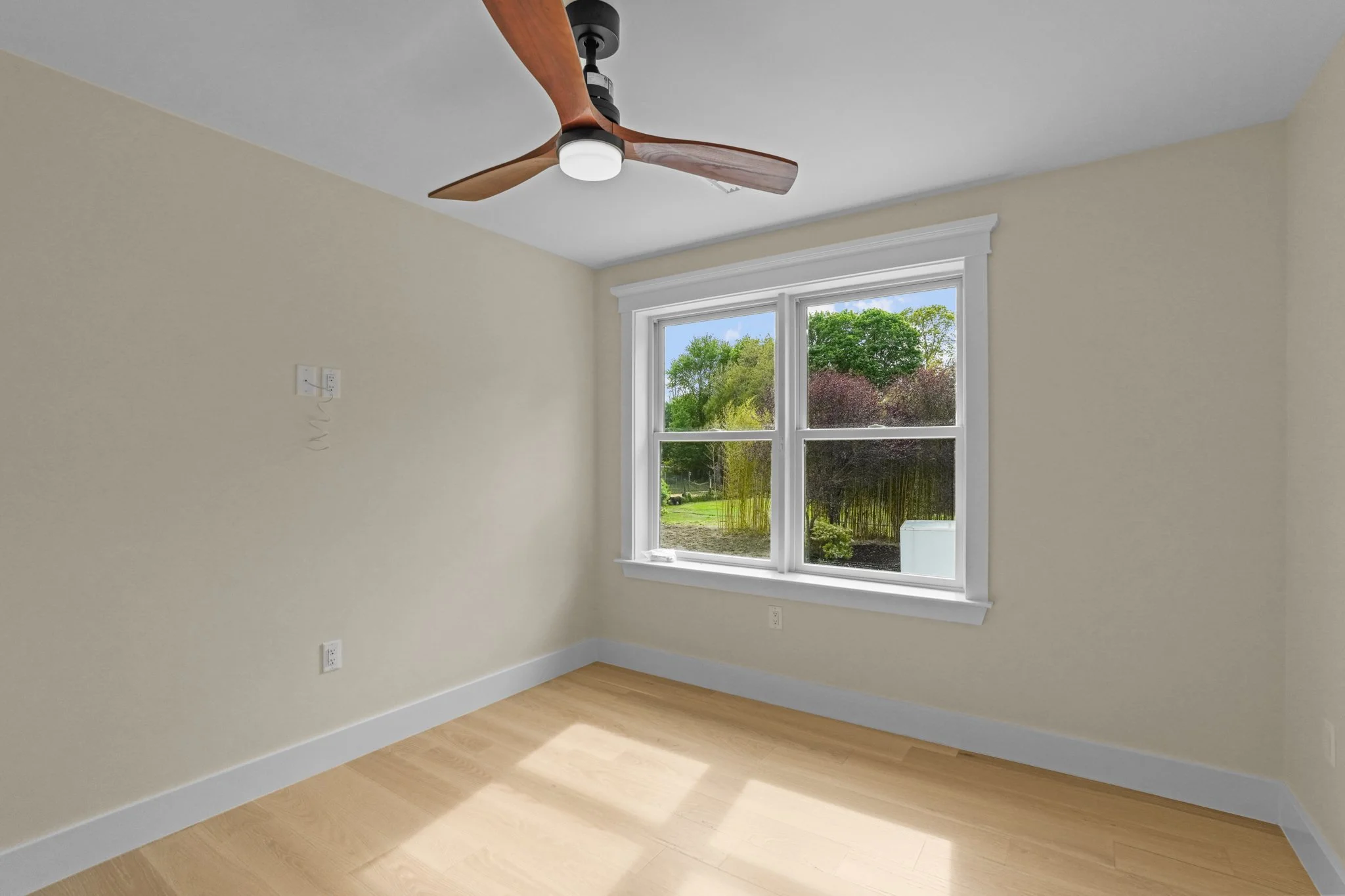 Empty room with beige walls, hardwood flooring, a white-framed window showing trees outside, a ceiling fan with wooden blades and a central light fixture.