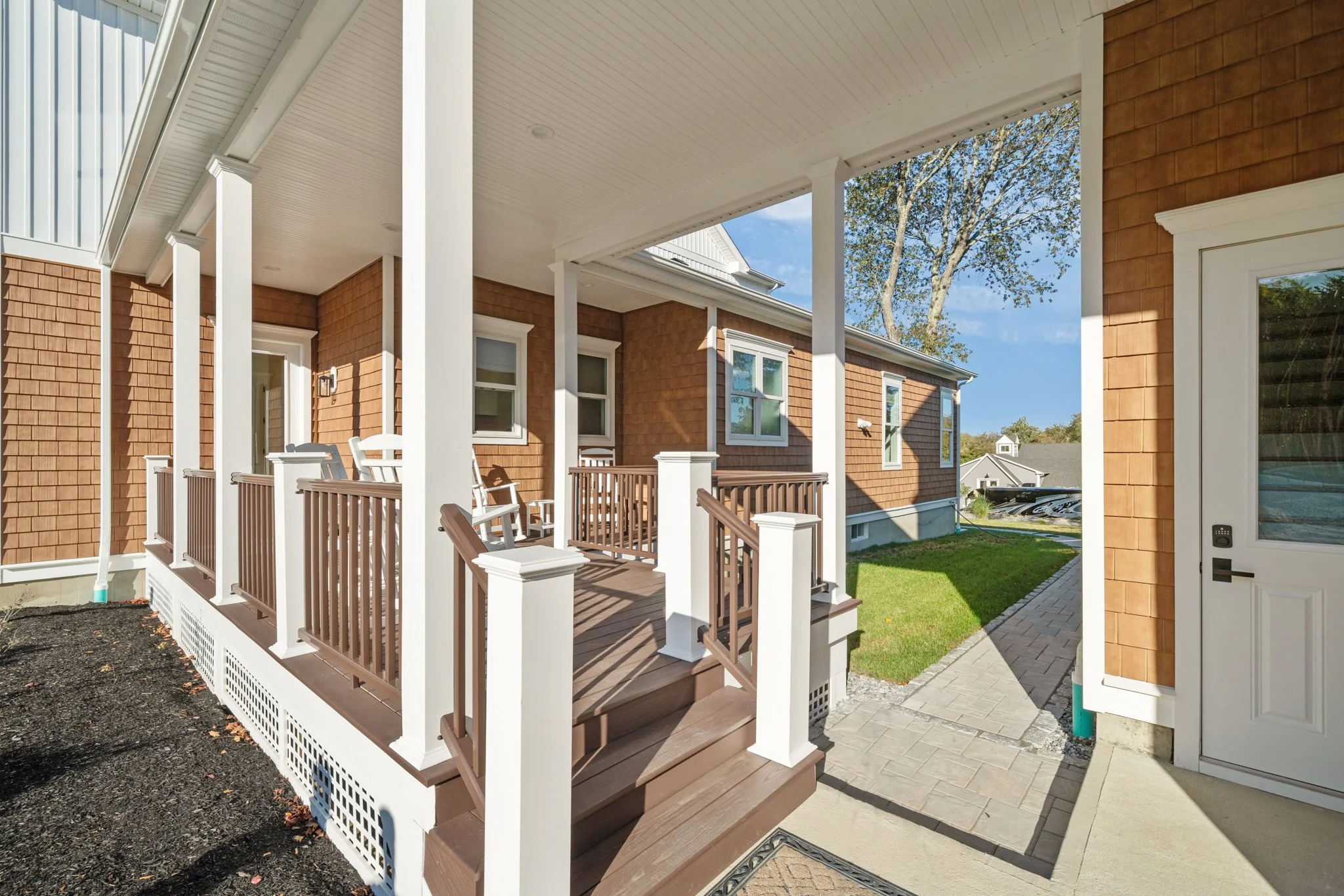 A front porch with wooden flooring, white railings, and columns attached to a house with brown shingle siding. There are white framed windows, a glass door, and a small garden area with a tree and neighboring houses in the background.
