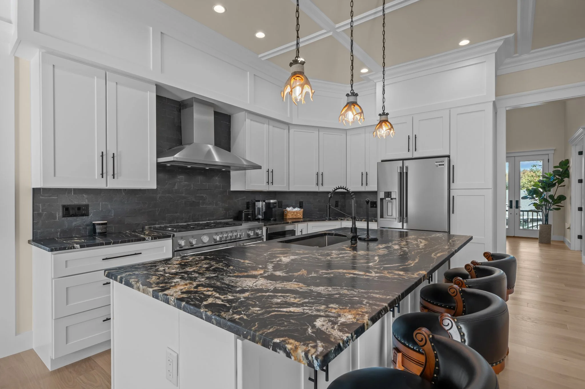 Modern kitchen with white cabinets, black and gold marble island, black backsplash, stainless steel appliances, decorative pendant lights, and a potted plant near a glass door.