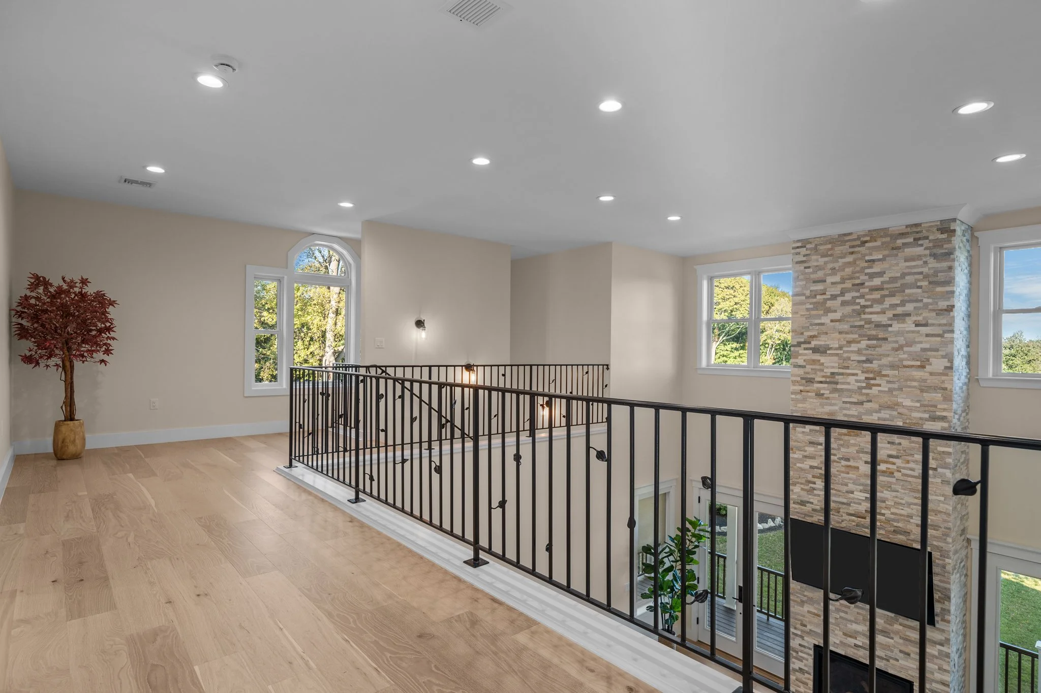 Interior view of a modern house with a balcony overlooking a staircase, featuring large windows, a brick accent wall, hardwood flooring, and a potted plant.