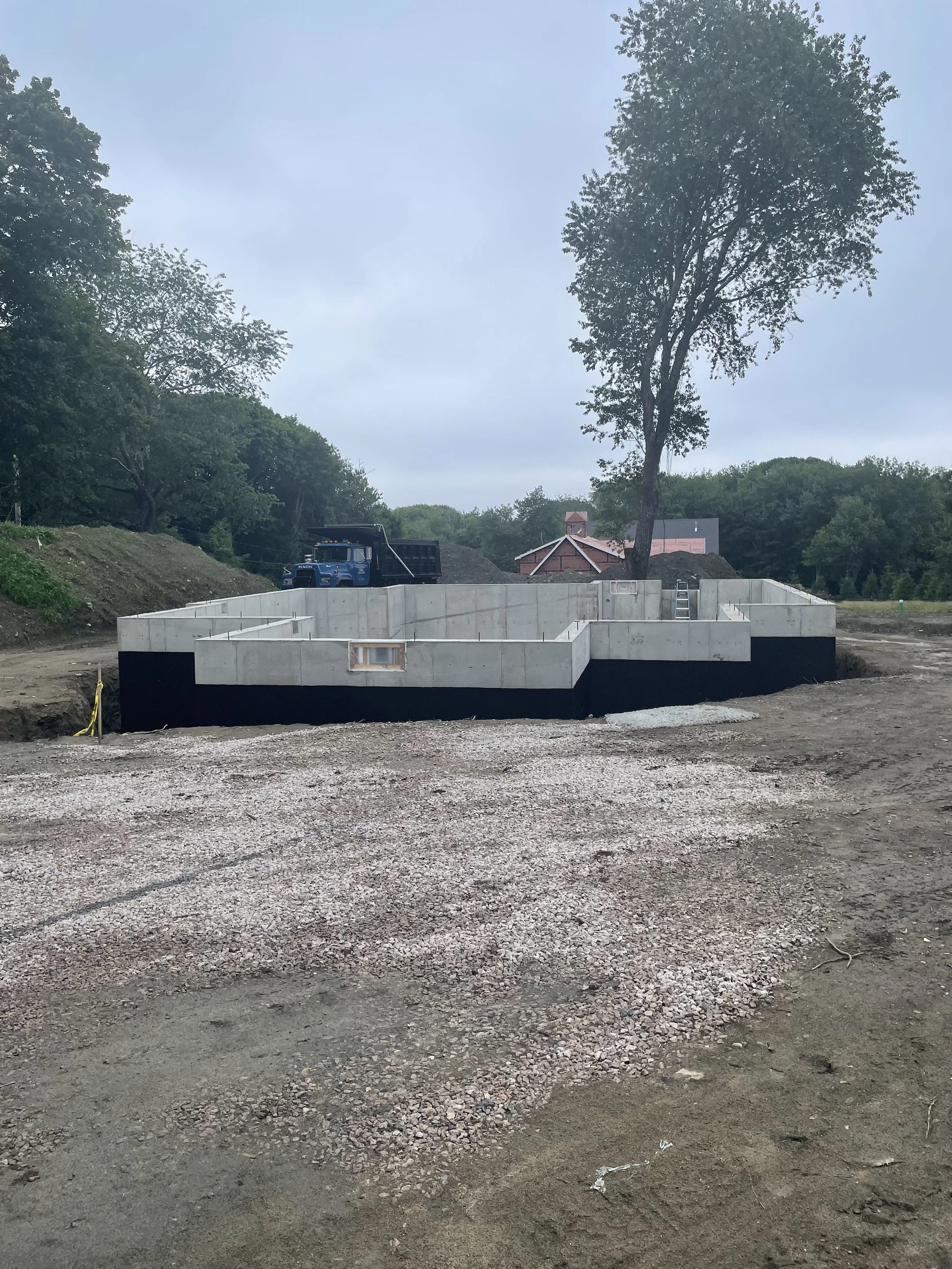 Construction site with concrete foundation, dirt, and gravel, surrounded by trees and cloudy sky.