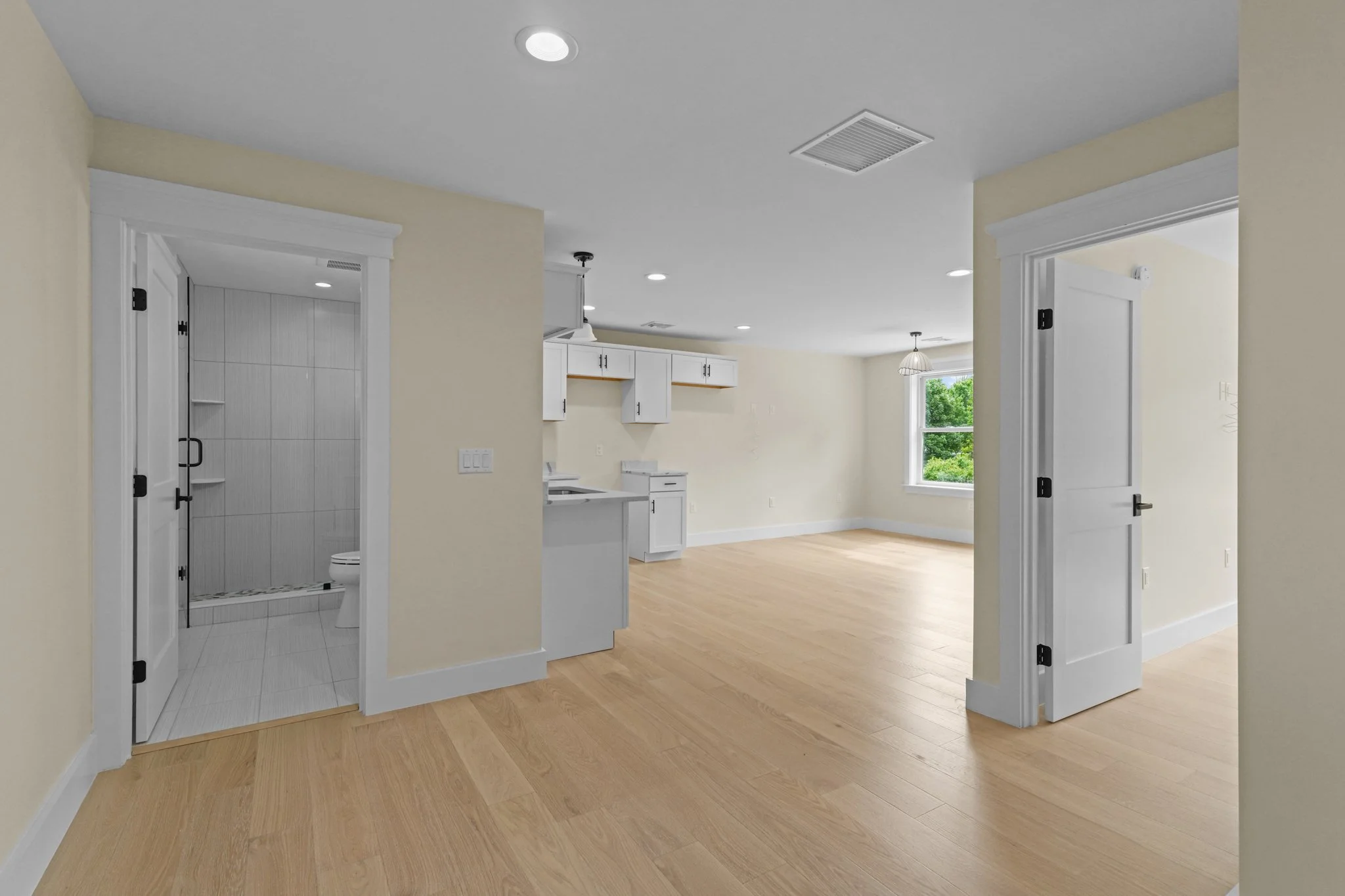 Empty living space with light wood floors, white trim and ceiling, beige walls, and white kitchen cabinets. There are two doorways with white trim, a window with greenery outside, and a small bathroom with a shower visible through the left doorway.