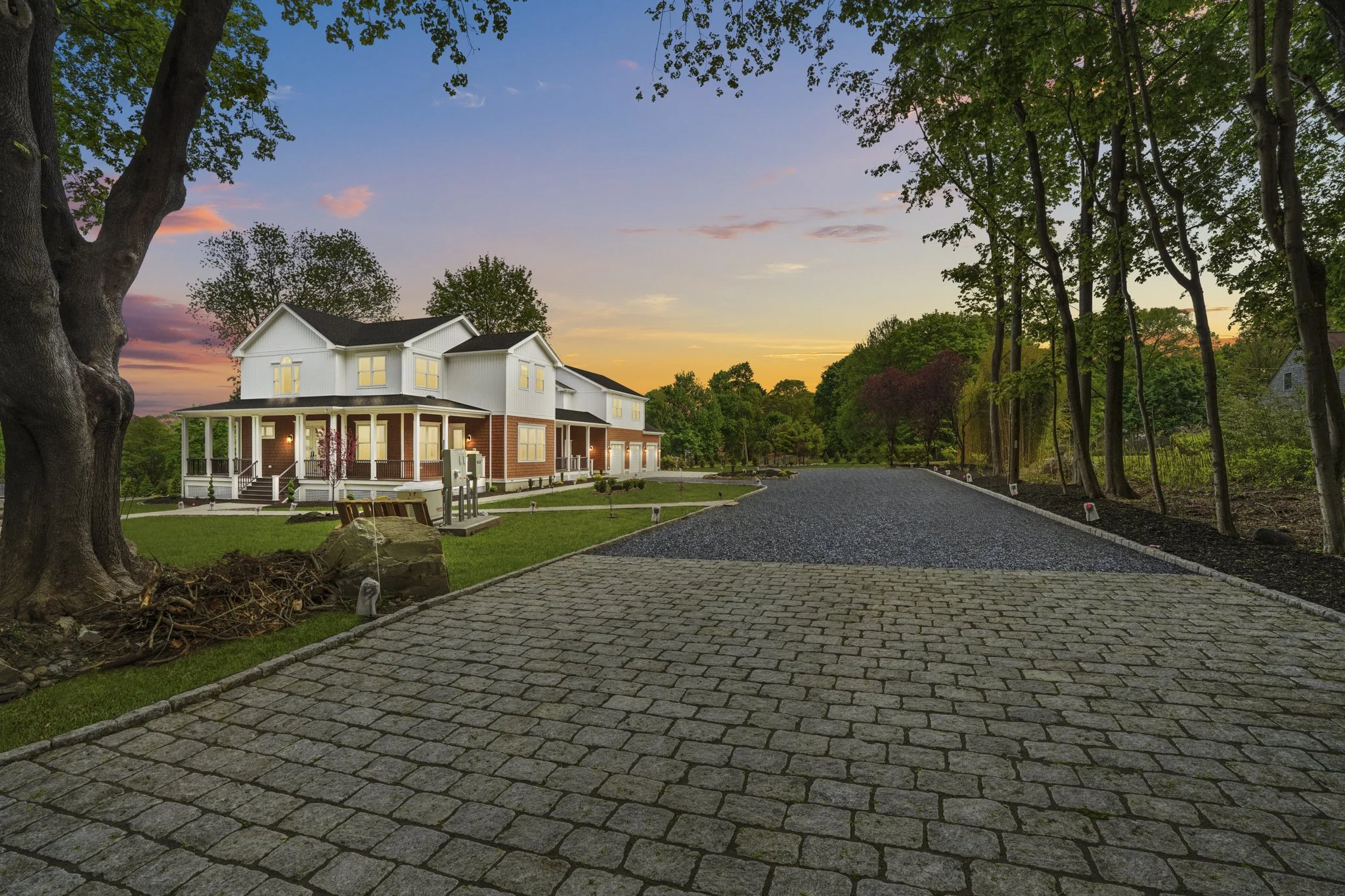 A large white house with a brick foundation and a wraparound porch, set in a wooded area. The driveway is paved with gray interlocking stones, leading up to the house. The sky is during sunset with pink, purple, and orange hues. Tall trees with green