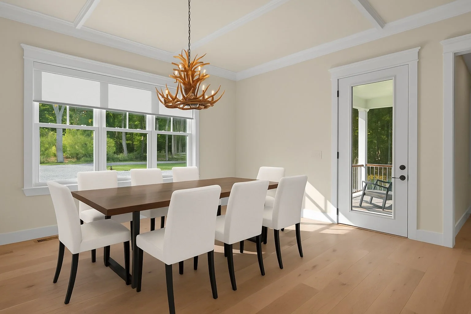Dining room with white chairs around a wooden table, large window showing trees outside, and glass door leading to a balcony with chairs.