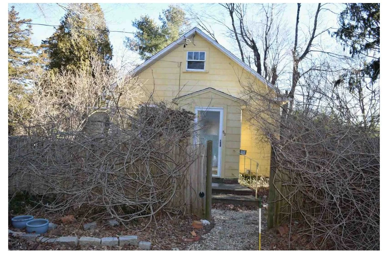 Yellow house with stairs leading to front door, surrounded by leafless bushes and trees, with a gravel pathway and a mailbox near the entrance.
