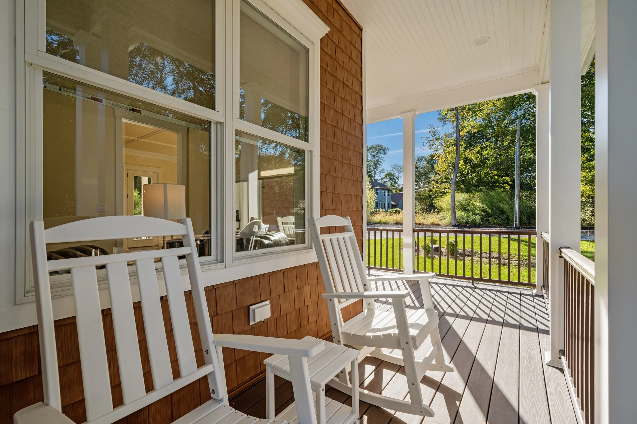 View of a house's front porch with white rocking chairs, wooden siding, large windows, and trees in the background on a sunny day.