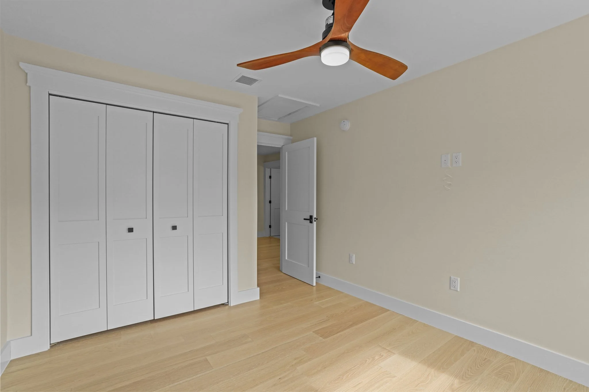 A room with a ceiling fan, cream-colored walls, and a closed white closet with sliding doors. The room features light wood flooring and an open door leading to another room.