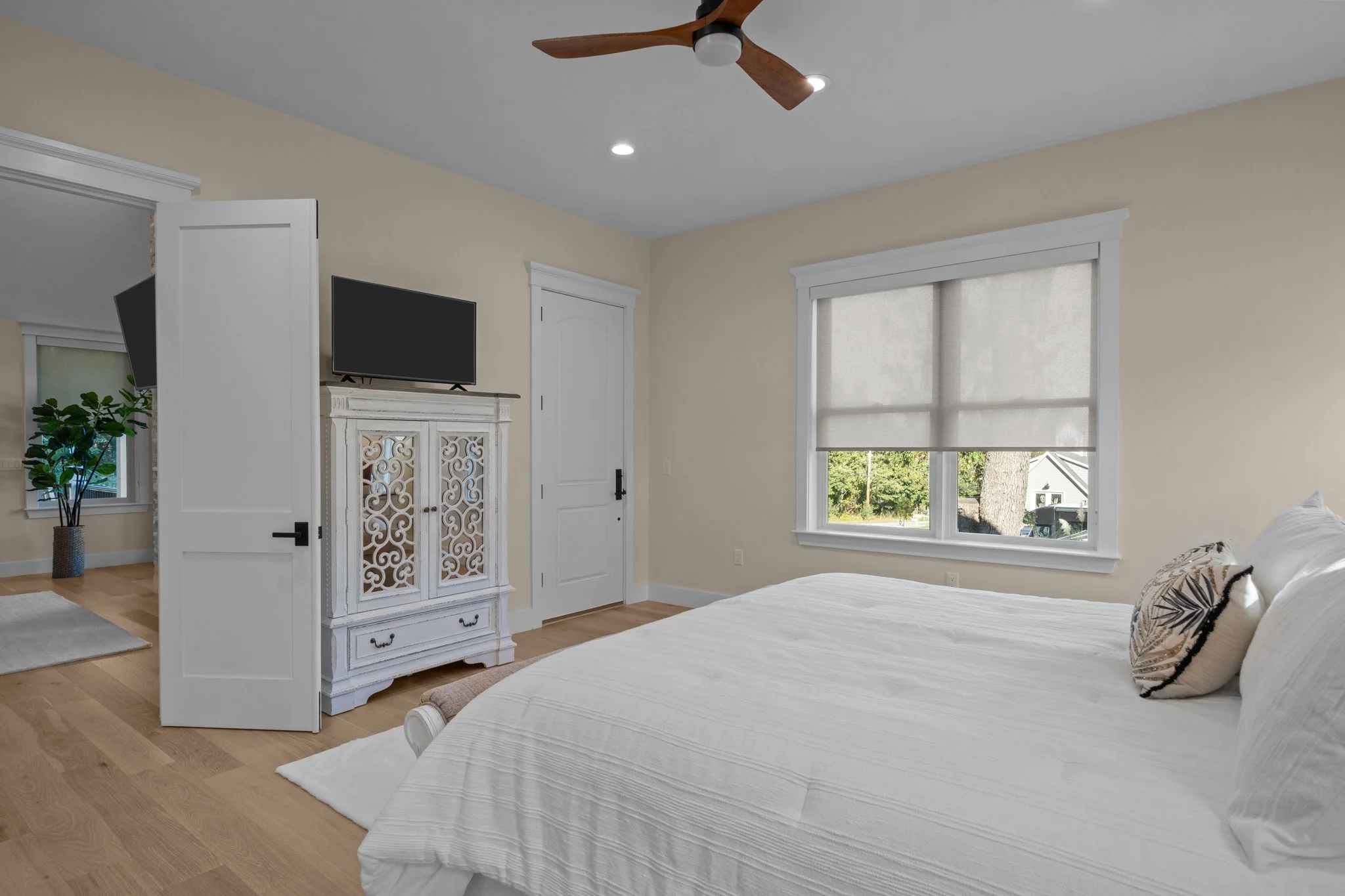 A bedroom with beige walls, white trim, a large window with blinds, and a white bed with a decorative pillow. There is a ceiling fan with wooden blades and a light, a flat-screen TV on a stand, and a white ornate cabinet. A small plant is in the corn