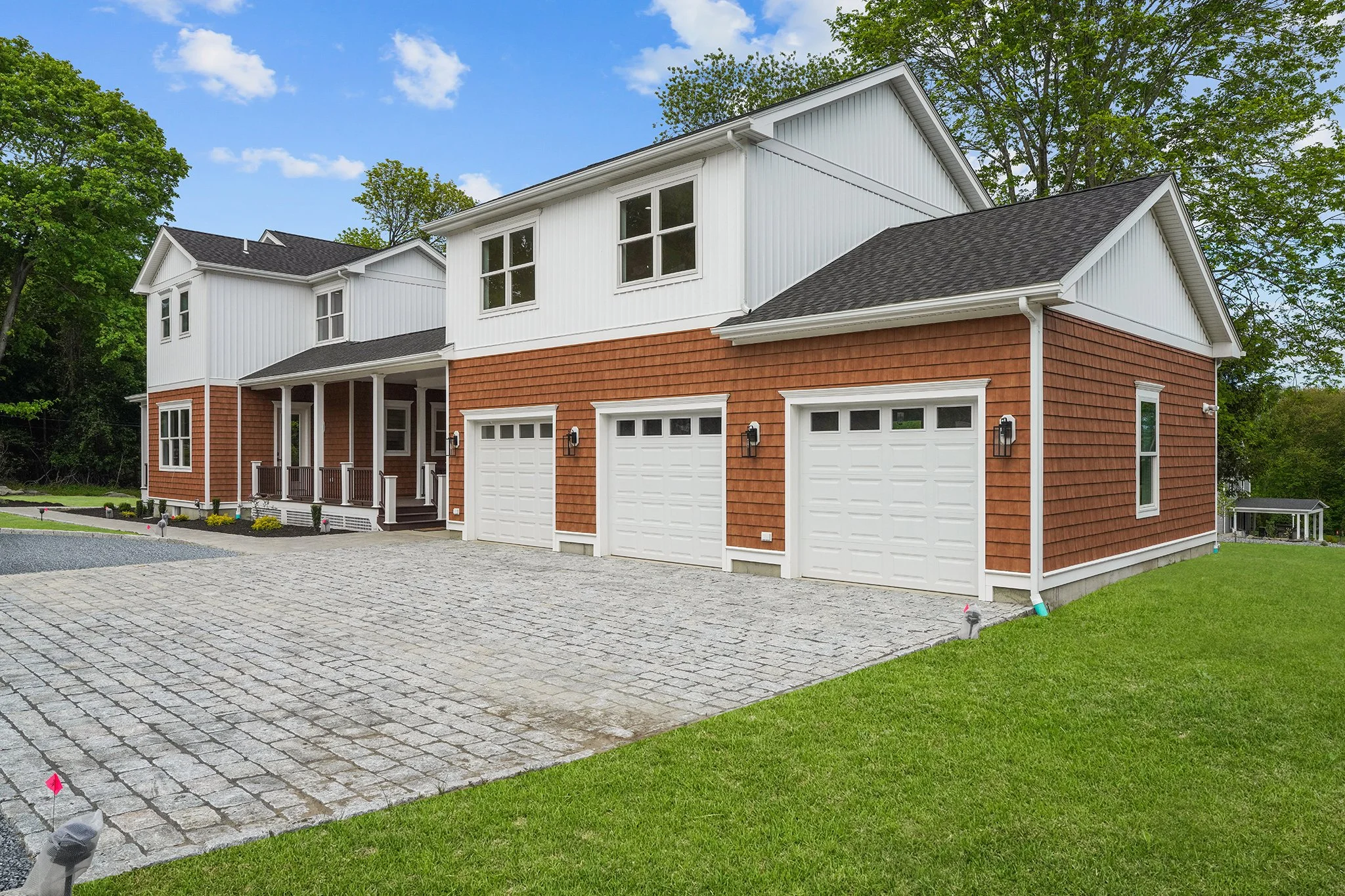 A two-story house with a combination of white siding and orange shingle exterior, black shingle roof, three garage doors, small front porch, and surrounded by green trees and grass.