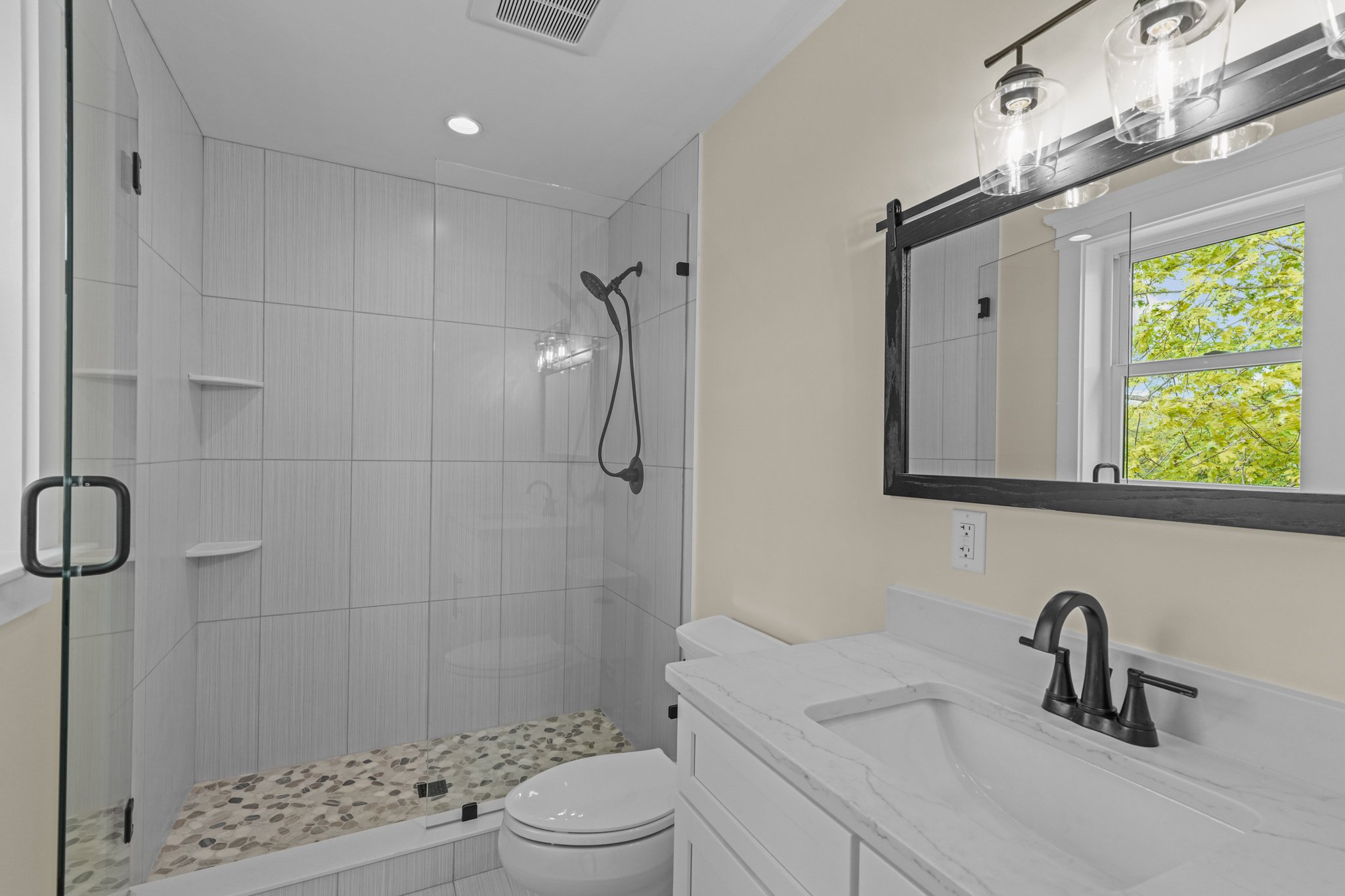 A modern bathroom with a walk-in shower with glass enclosure, a white vanity with a marble countertop and black fixtures, a window showing green trees outside, and ceiling lights.