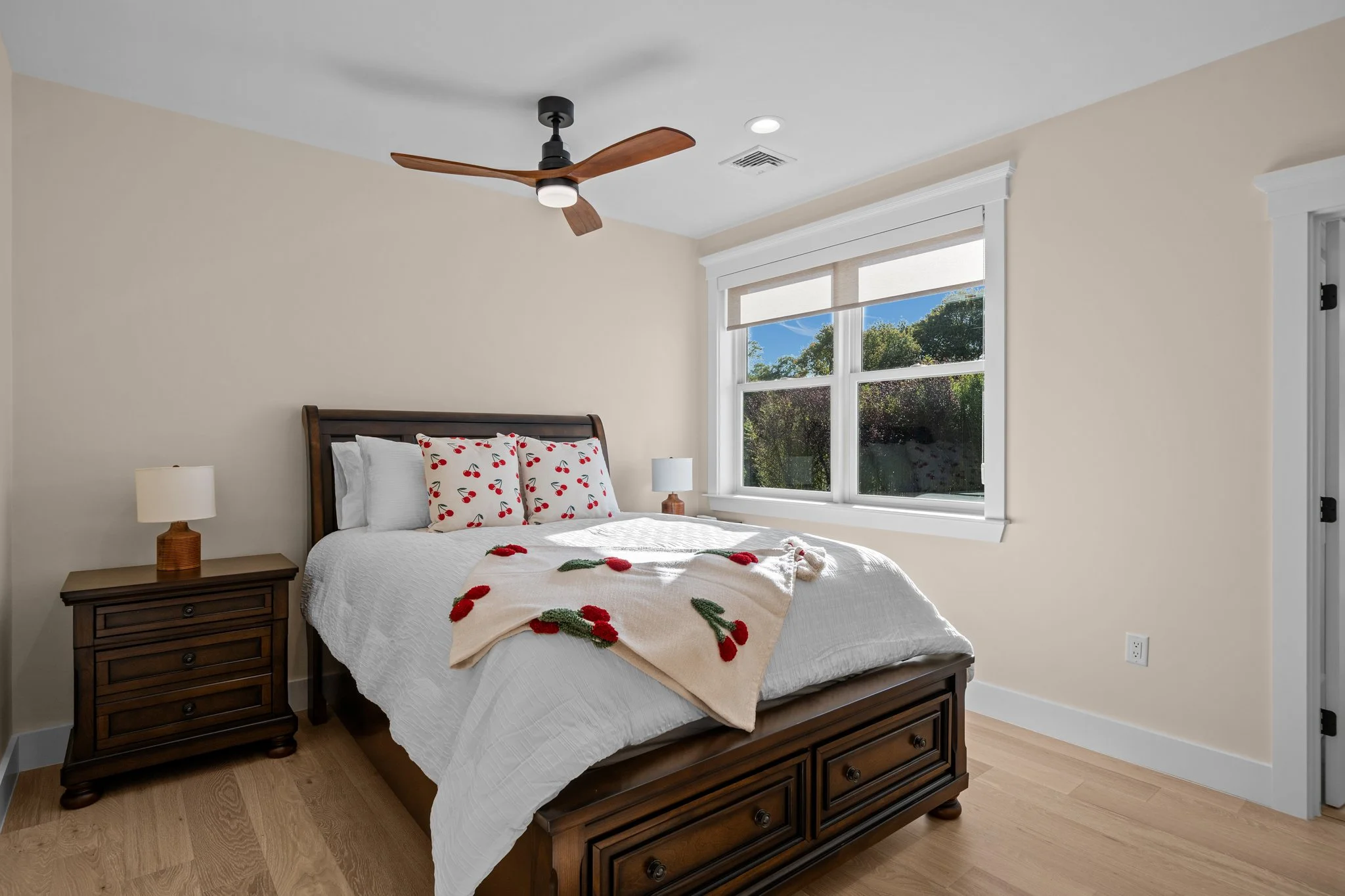 A cozy bedroom with a wooden bed frame, white bedding adorned with cherry patterns, and a Christmas-themed blanket. There are two matching nightstands with lamps, a large window with a view of trees outside, and a ceiling fan with wooden blades.