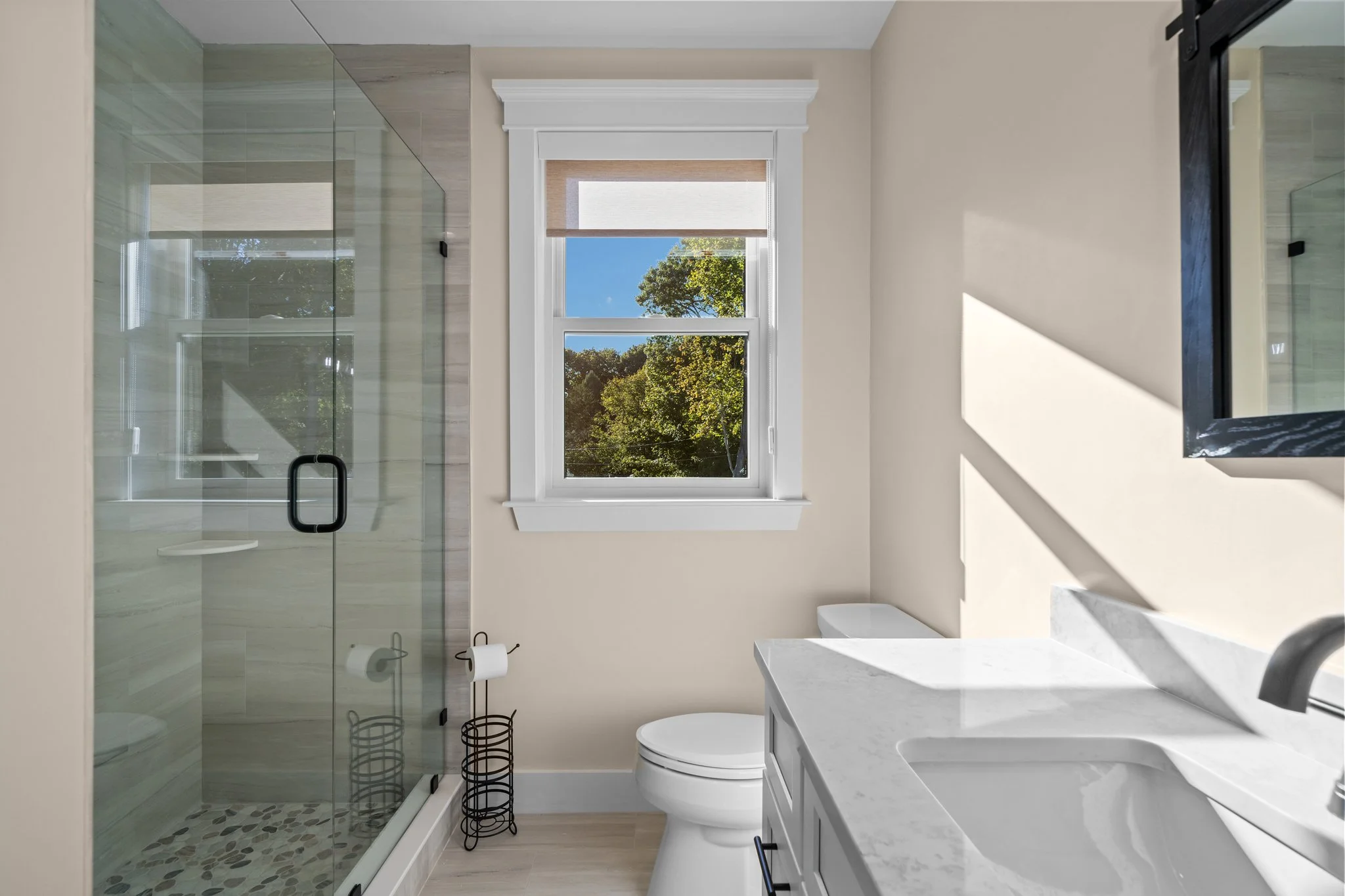 Bathroom with a glass shower enclosure, a toilet, a white vanity with a marble countertop, a wall-mounted mirror, and a window showing trees and a clear blue sky outside.