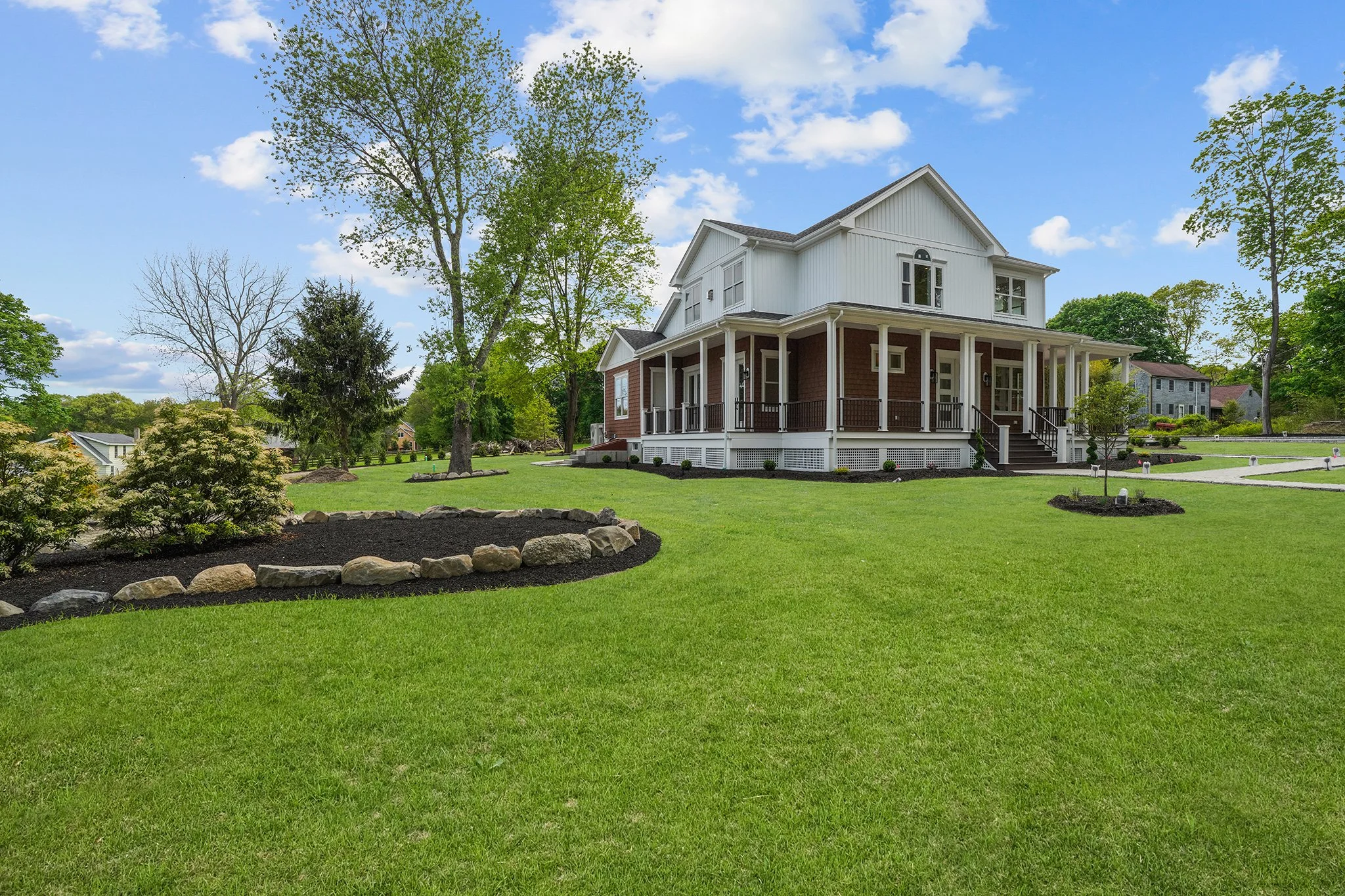 A large white two-story house with a front porch, surrounded by a green lawn, trees, and shrubs, under a blue sky with clouds.