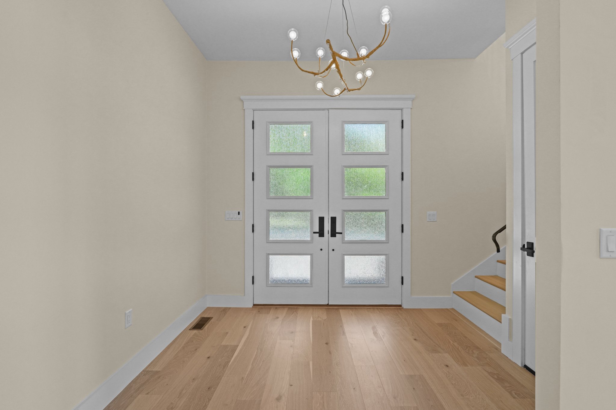 Entryway with double front door with glass panes, light-colored walls, hardwood floor, chandelier hanging from ceiling, staircase partially visible on the right.