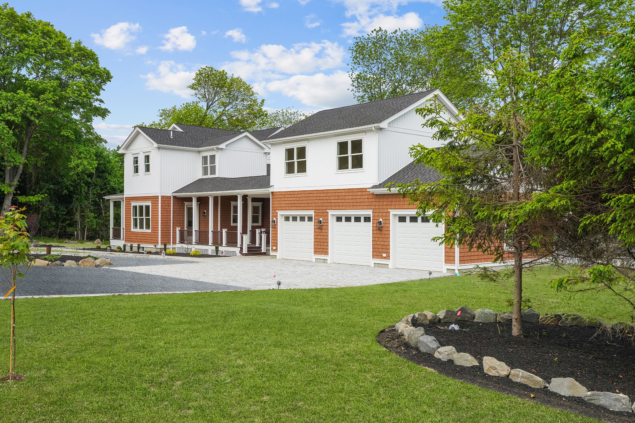 Front view of a two-story house with white siding and a brown shingle accent on the lower level, garage doors, a porch, green lawn, trees, and a partly cloudy sky.