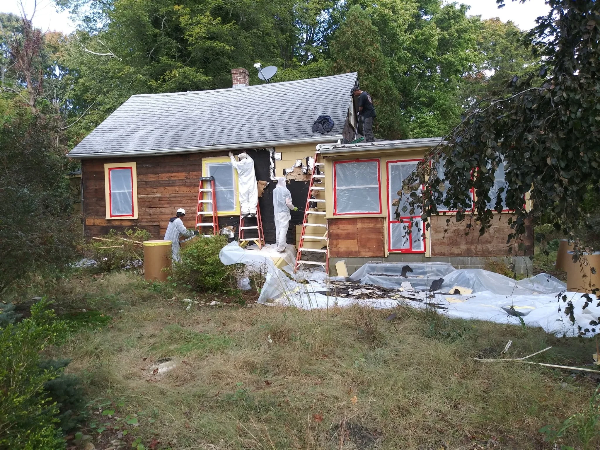 Construction workers repairing the exterior of a small house surrounded by trees. Some workers are on ladders, and tarp covers the ground.