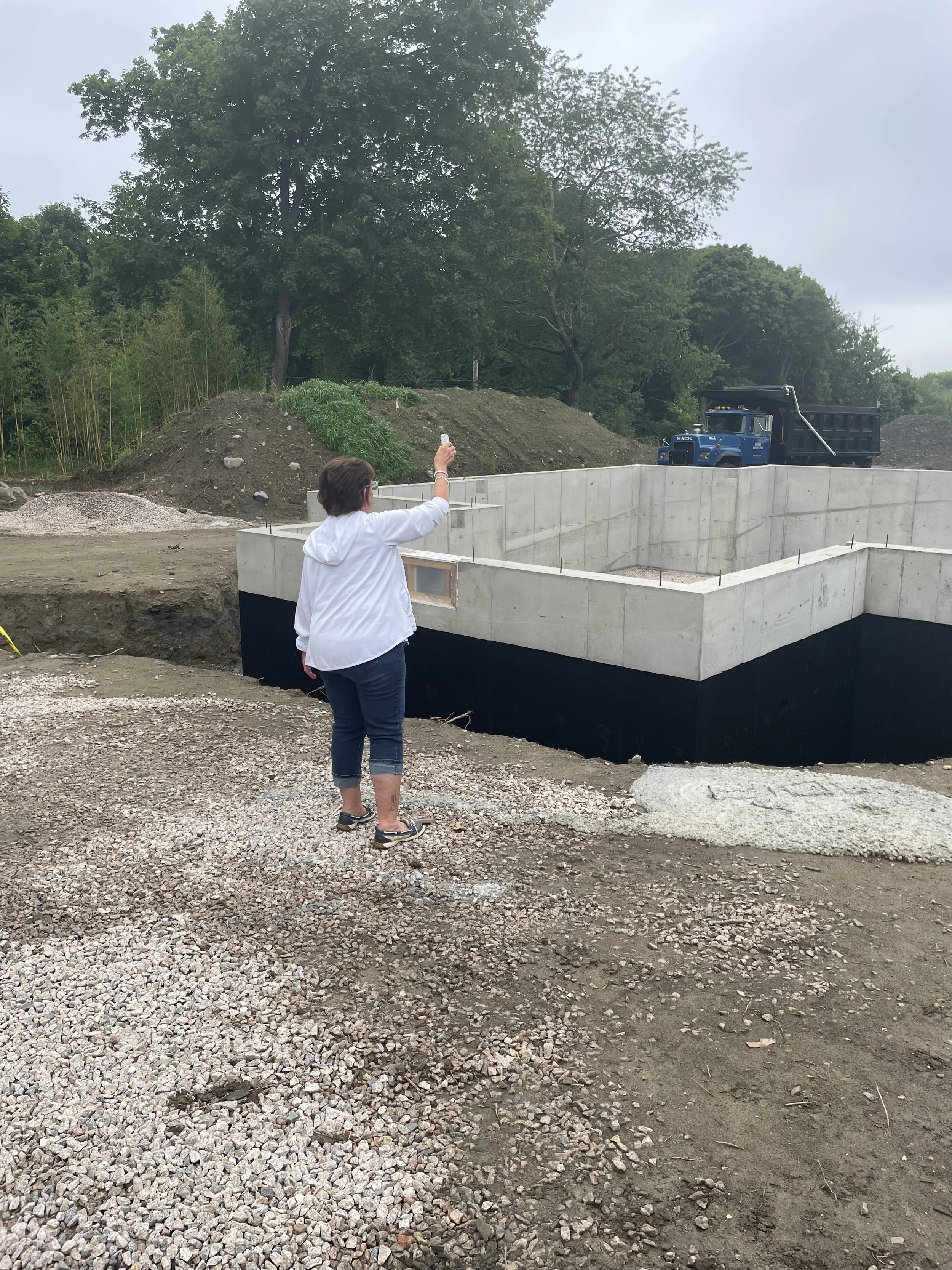 A woman with short dark hair wearing a white hoodie and jeans is standing on a gravel surface at a construction site, taking a photo with her phone of a concrete foundation and a blue flatbed truck in the background, with trees and an overcast sky ov