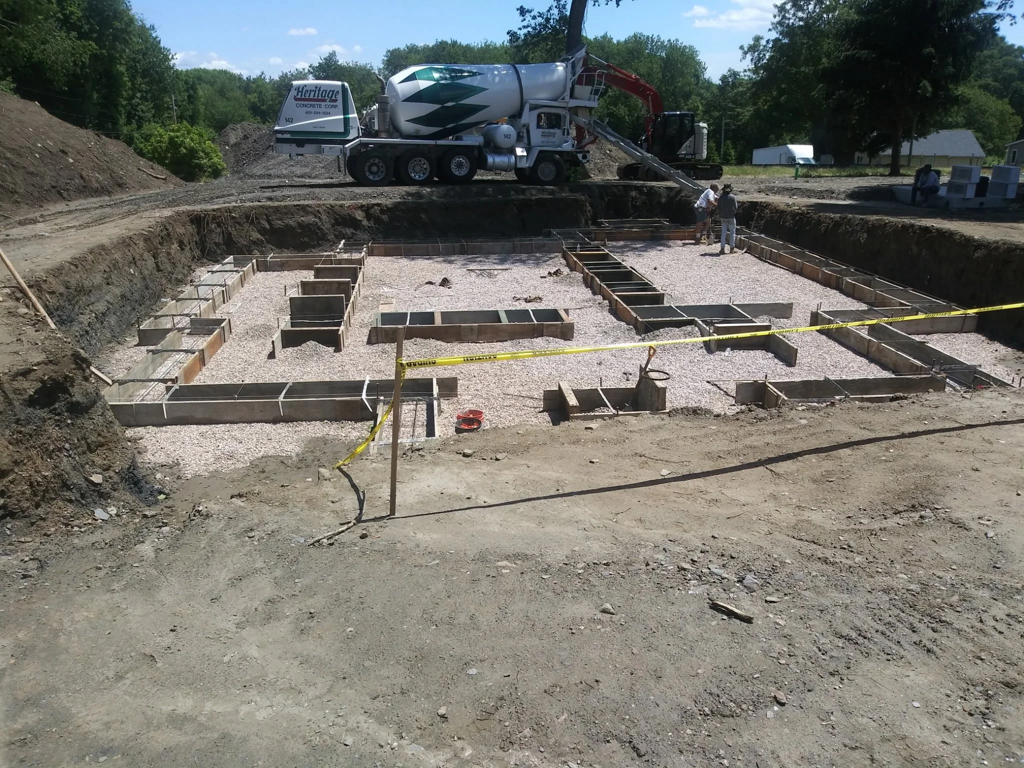 Construction site with a cement mixer truck pouring concrete into a foundation framework, three workers nearby, and trees in the background.