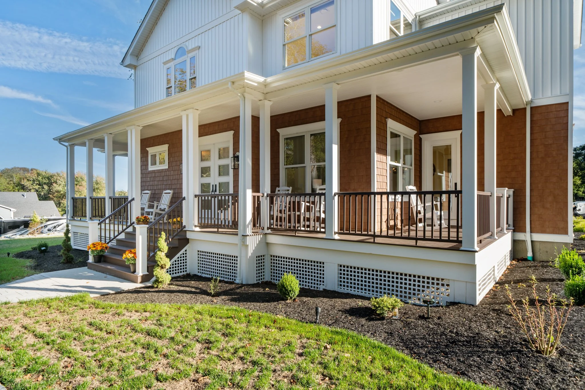 Front porch of a house with four white rocking chairs, potted plants, and a wooden railing. The house has white siding and brown brick sections, with large windows and a covered balcony.