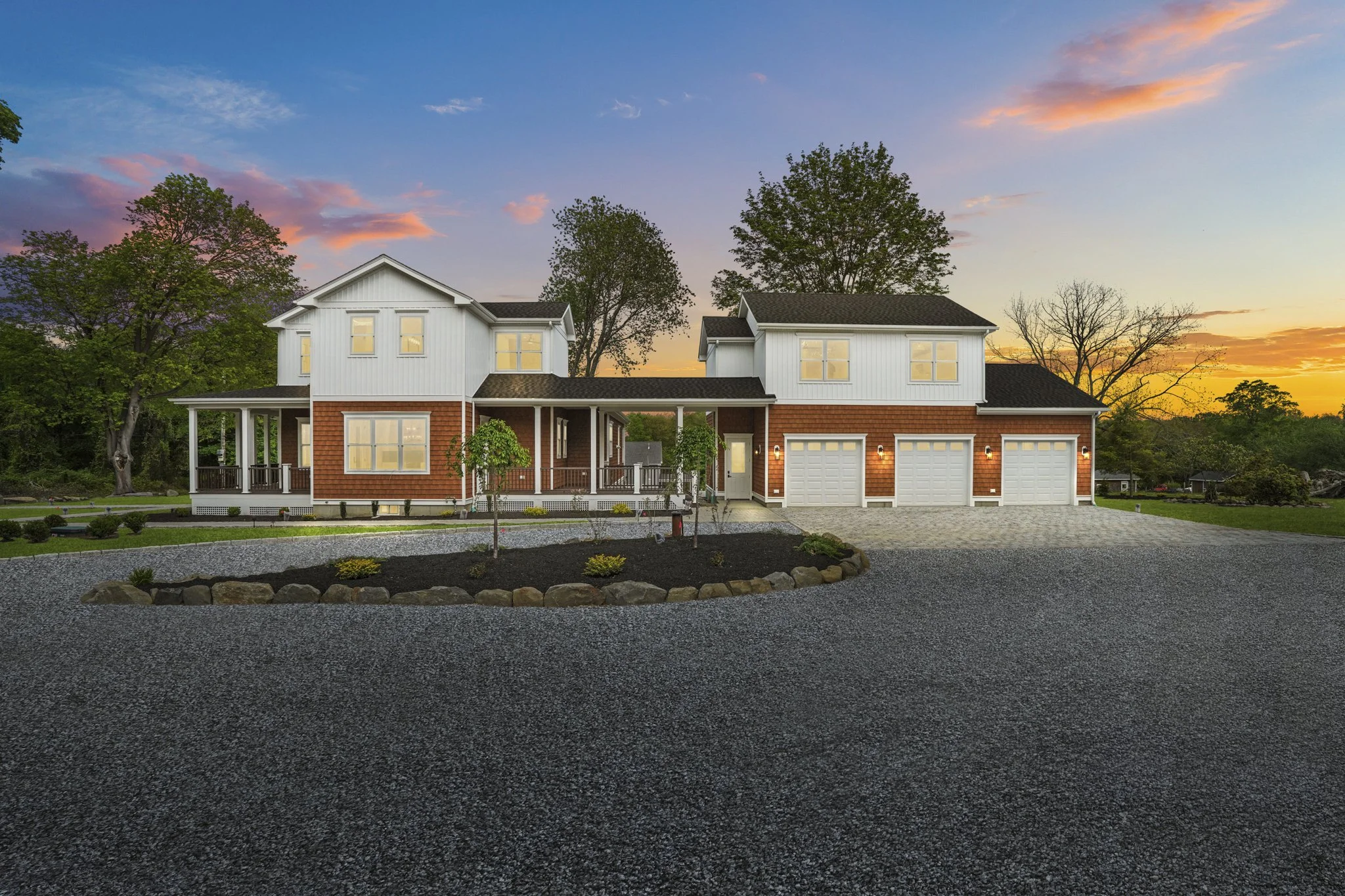 A two-story house with white and red siding, large windows, a covered porch, and a three-car garage, surrounded by trees and a landscaped yard at sunset.